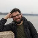 CryptoGain Google Play Store reviewer Alec Opperman profile photo showing bearded man with glasses smiling outdoors by waterfront wearing casual black jacket