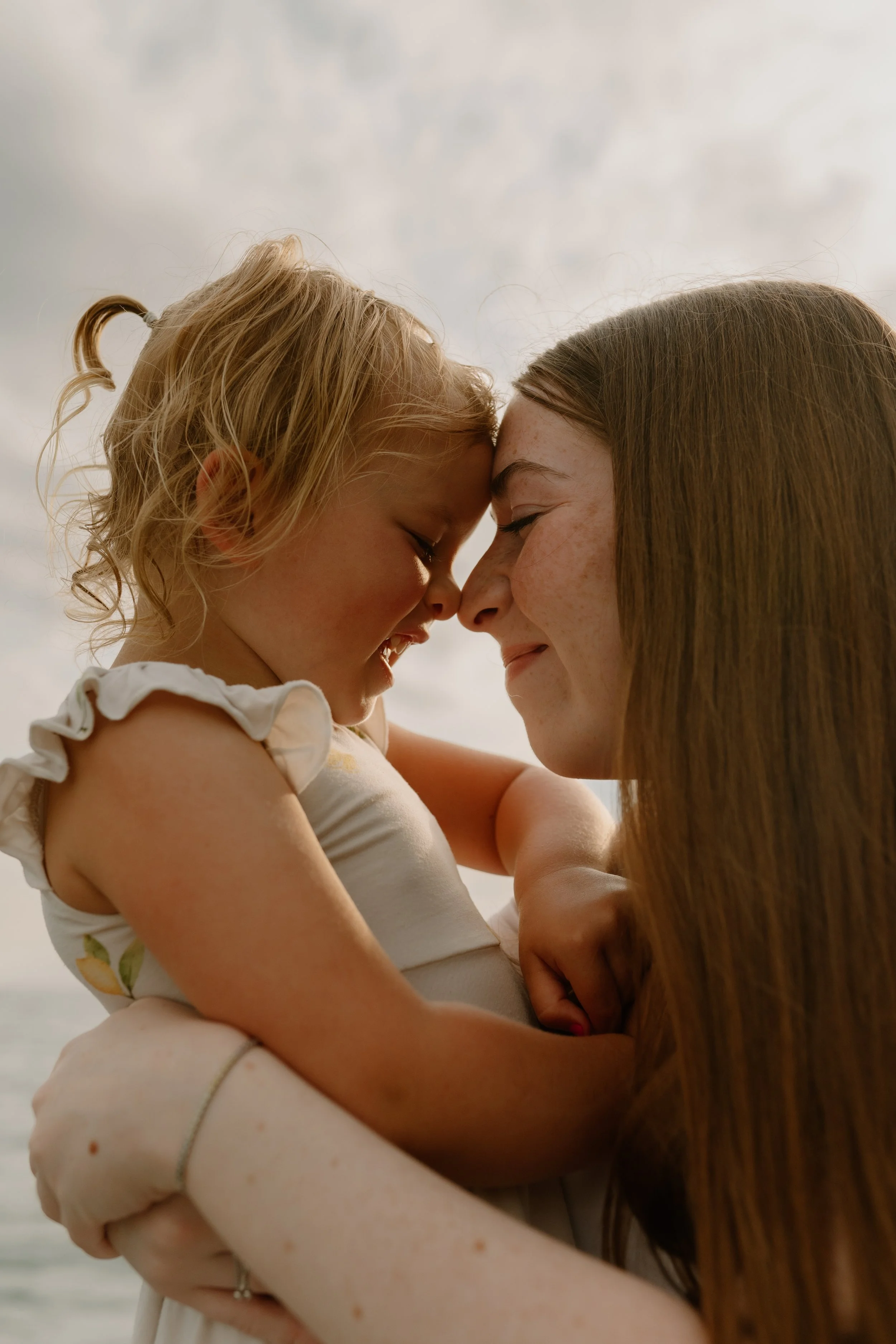 A woman with long brown hair wearing a cream crocheted top holding a blonde curly-haired child on a hammock in a wooded area, with a lake in the background, during their Ontario family photo session.