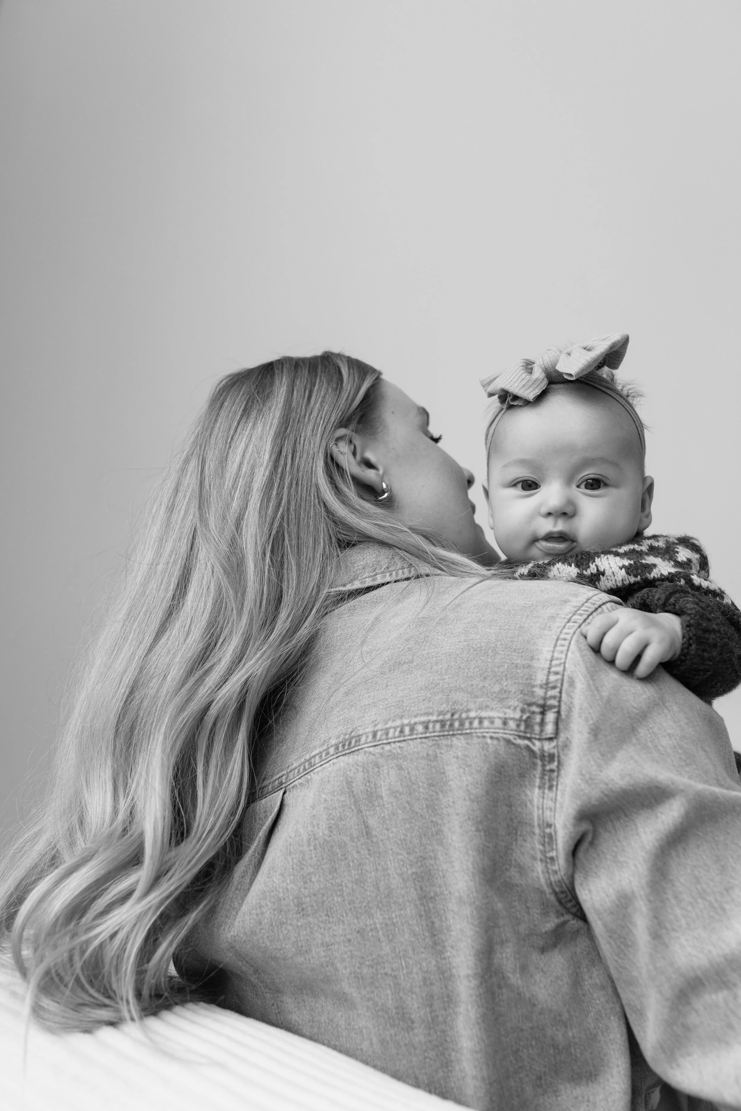 A woman with long wavy hair holding a baby wearing a large bow headband. The woman is facing left and the baby is looking directly at the camera. The background is plain and neutral.