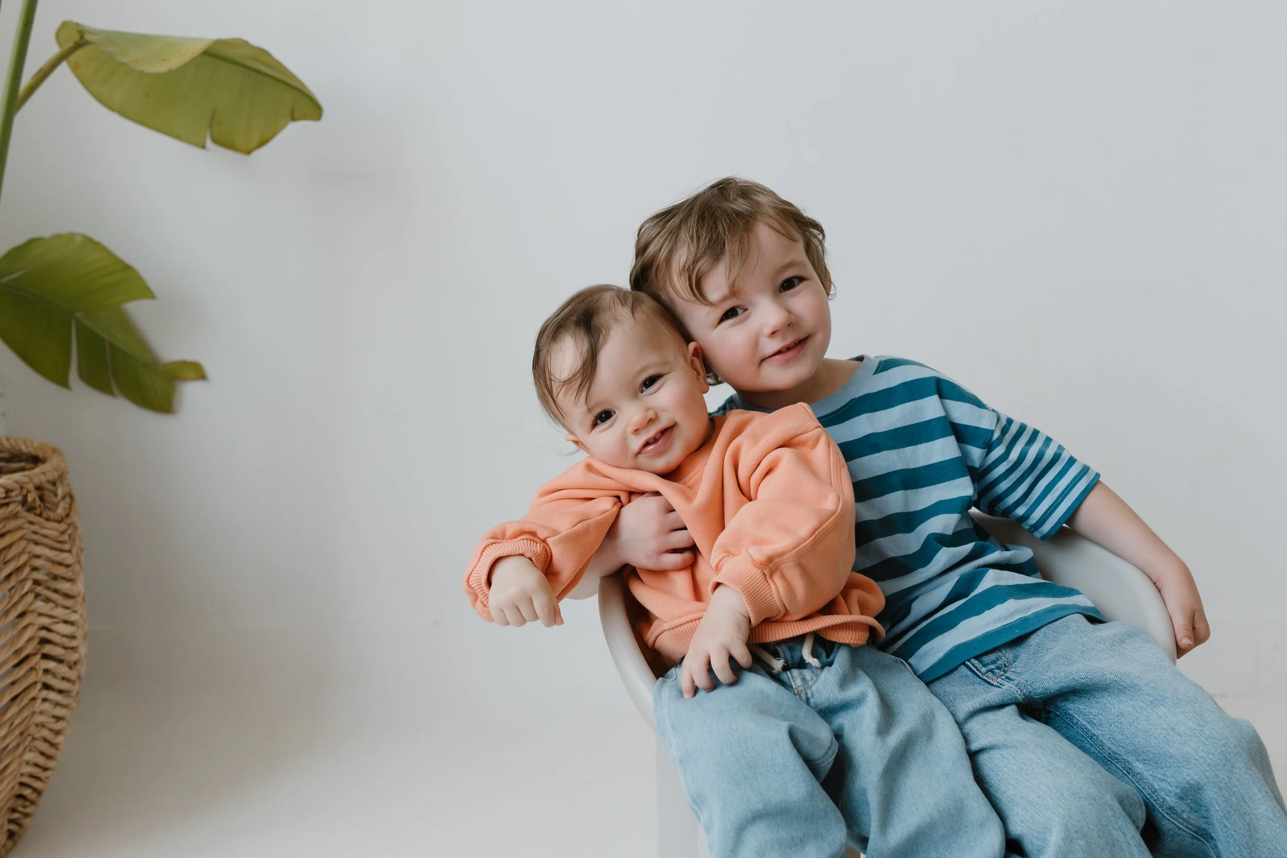 Two young boys sitting together, smiling, with the older boy holding the younger one in front of a plain white wall in a studio, with a large green leaf from a houseplant to the left, during their family photo session in Whitby Ontario.