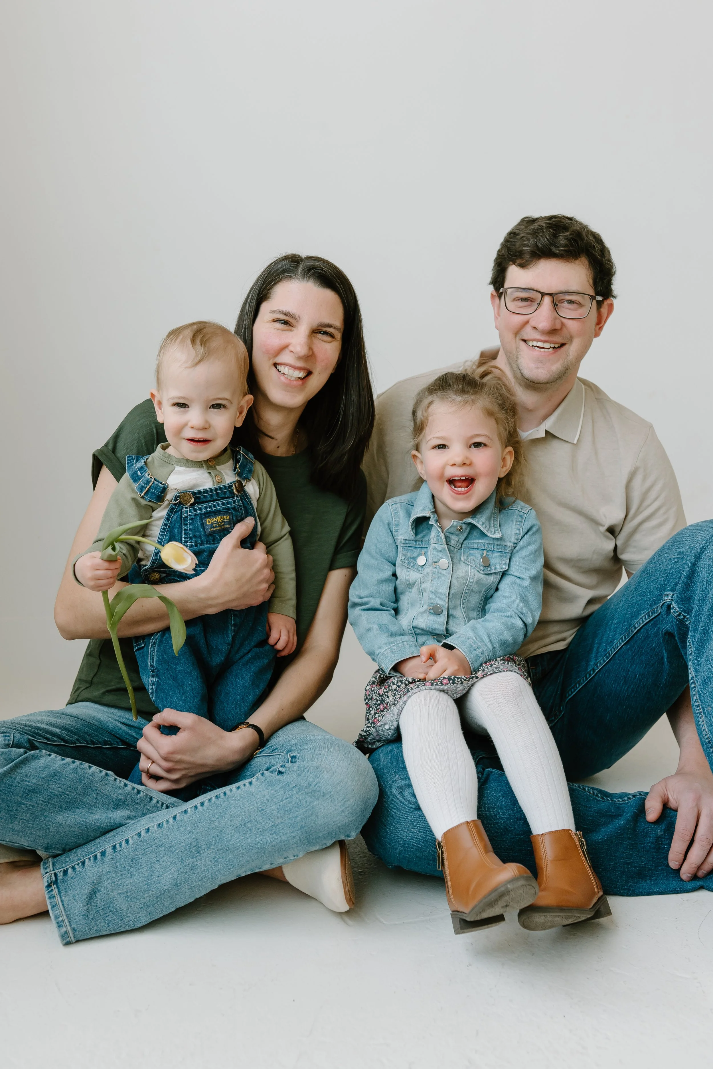 A happy family of four sitting on the floor against a plain white wall, smiling at the camera. The mother has dark hair, and the father has glasses. The children are a young boy holding a flower and a young girl with curly hair, both smiling.