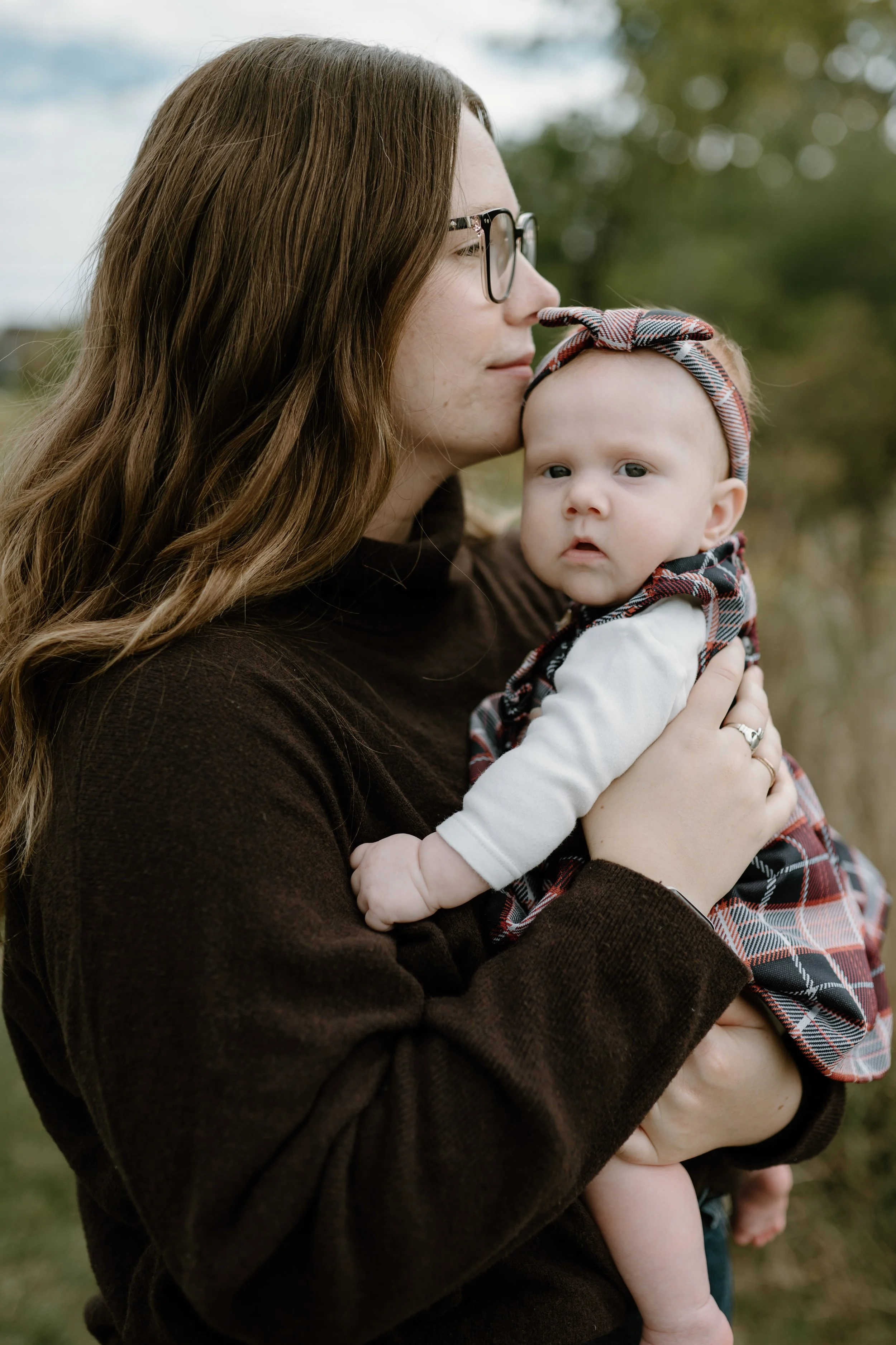 A woman with long brown hair, glasses, and a dark brown sweater holding a baby girl outdoors. The baby has light skin, blue eyes, a plaid headband, and a plaid dress. The woman leans in to kiss the baby on the forehead. Blurred trees and sky are in the background.