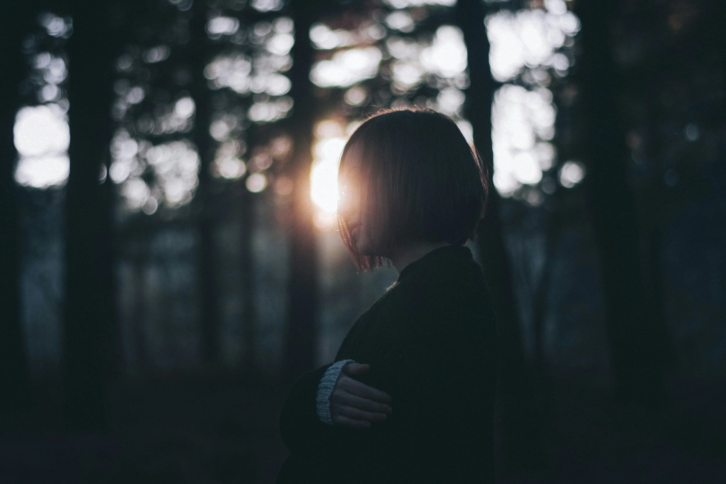 A woman with shoulder-length hair standing in a dark forest with tall trees, with the sun setting behind her, casting a warm glow around her head.