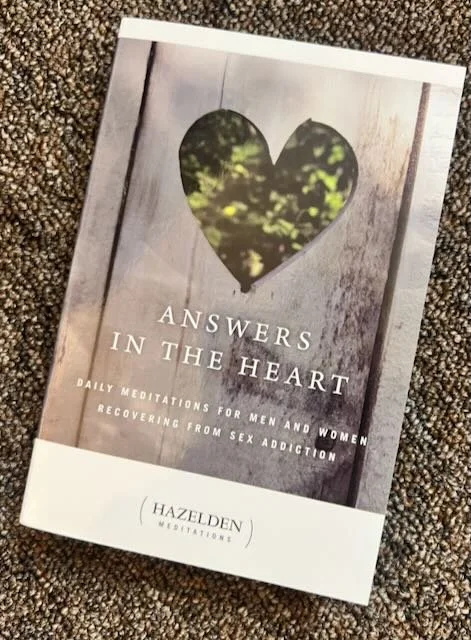 Book titled "Answers in the Heart" with a heart-shaped window showing greenery on a wooden background, about daily meditations for men and women recovering from sex addiction.