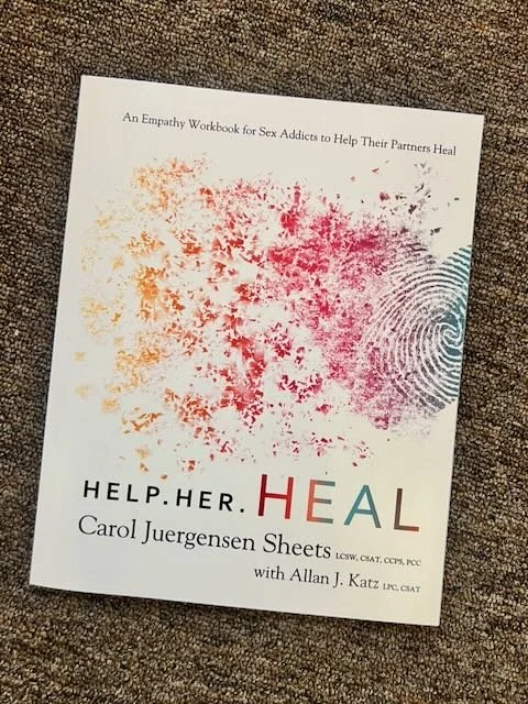 Book titled 'Help. Her. Heal.' by Carol Juergensens Sheets with a fingerprint and colorful splatter design on the cover, placed on a brown textured surface.