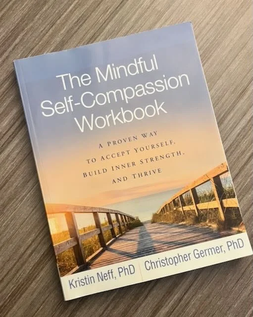 The cover of a book titled "The Mindful Self-Compassion Workbook" with a subtitle "A proven way to accept yourself, build inner strength, and thrive". The authors are Kristin Neff, PhD, and Christopher Germer, PhD. The cover features an image of a wooden boardwalk leading into the distance during sunset or sunrise.