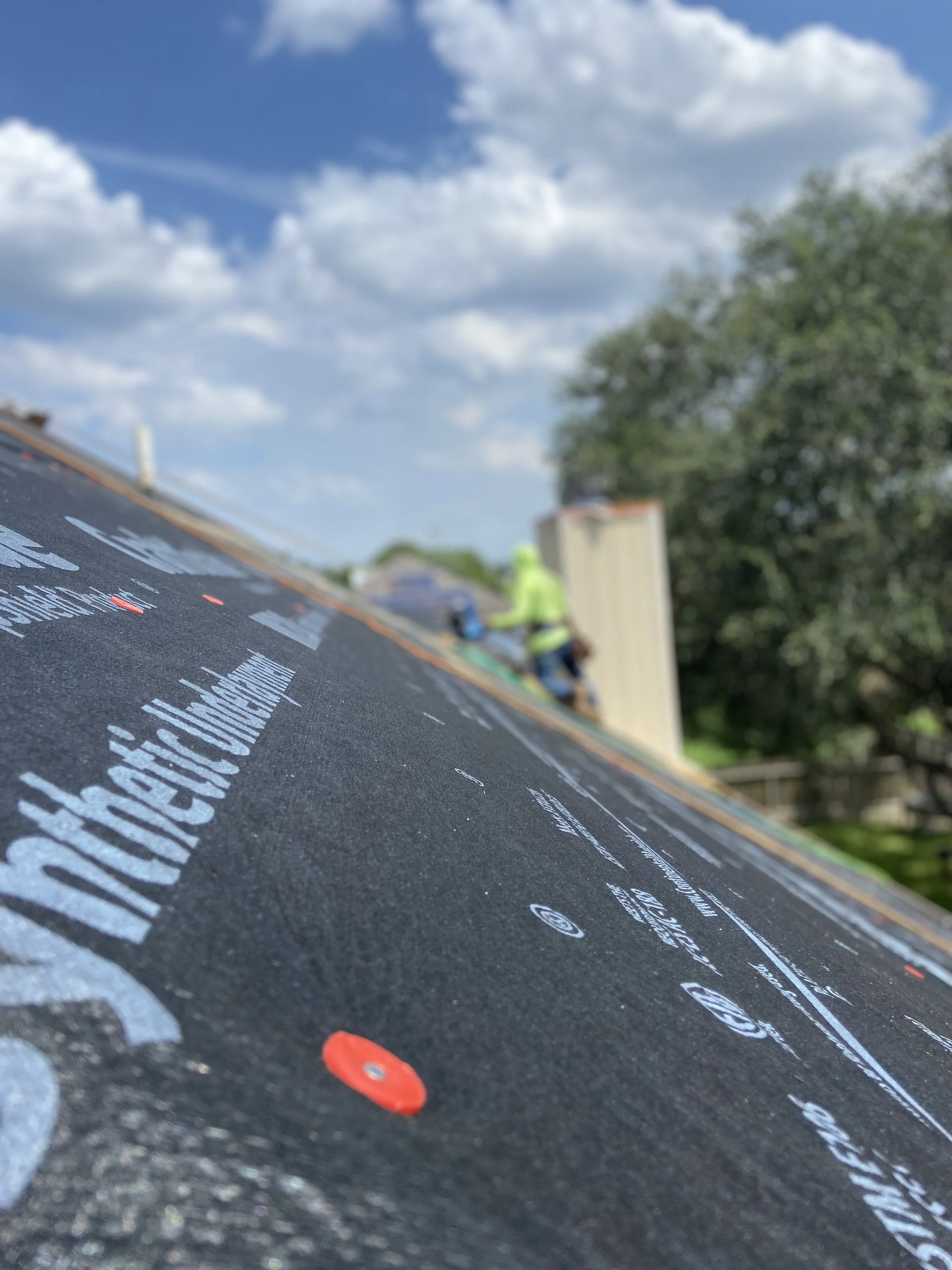 Close-up of a black roofing underlayment with red fasteners, with two workers installing a roof in the background under a blue sky with clouds.