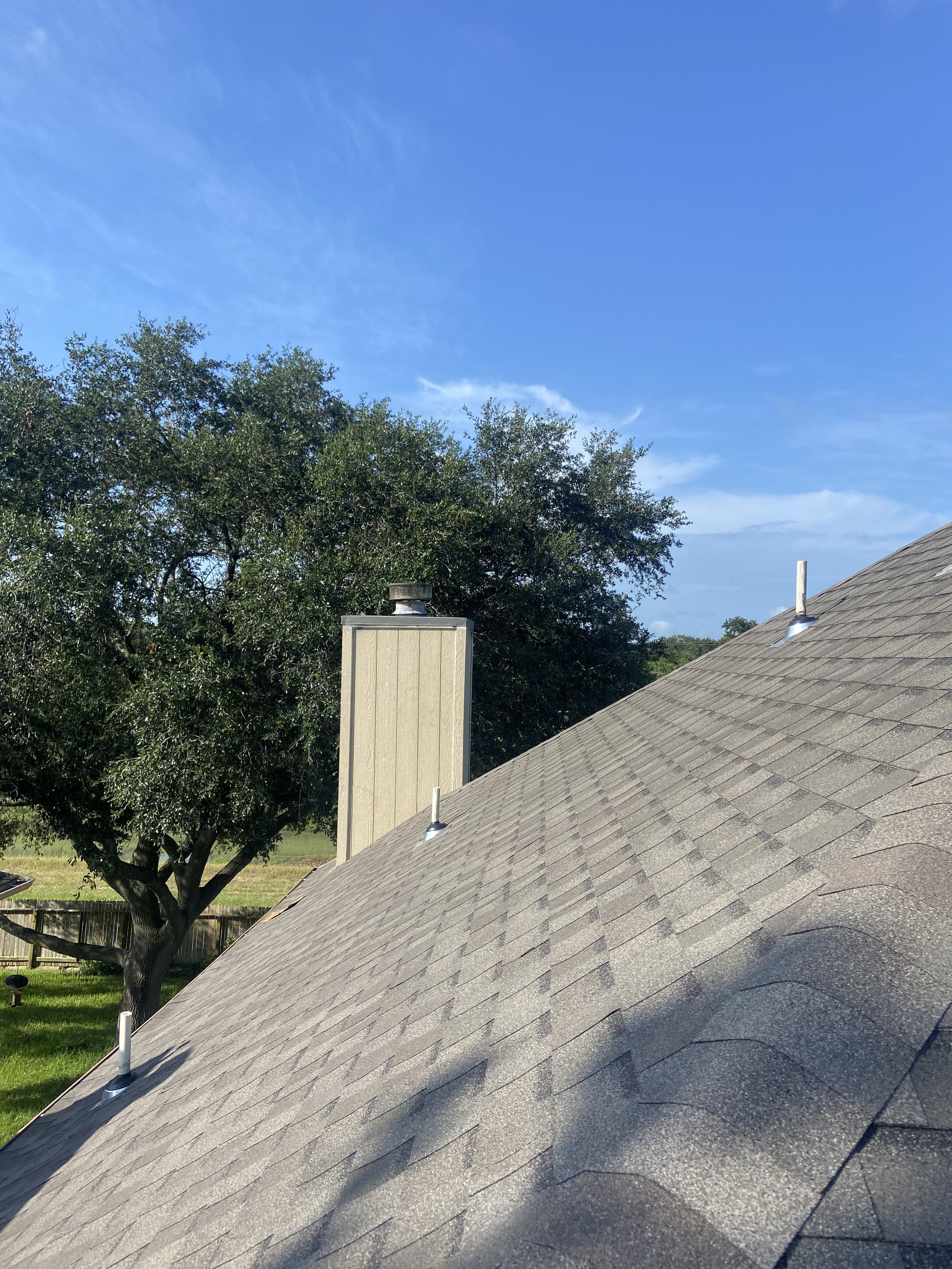 View of a house roof with asphalt shingles, two roof vents, a chimney, a large tree in the yard, and a clear blue sky.