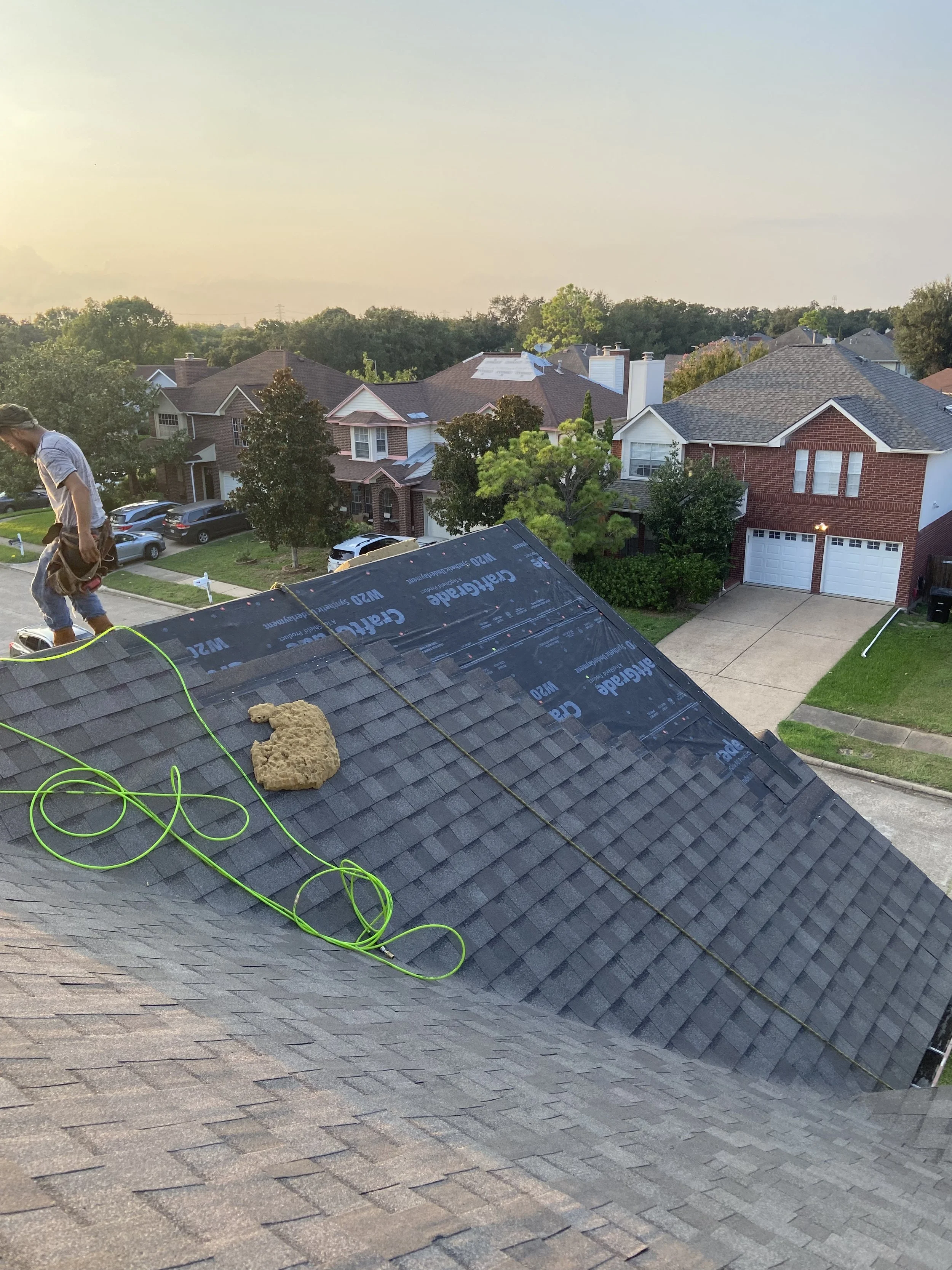 Roof of a house under renovation with some old shingles removed and new underlayment visible. A worker is standing on the roof near the edge, with tools and materials nearby, overlooking a neighborhood with other houses, trees, and a driveway.