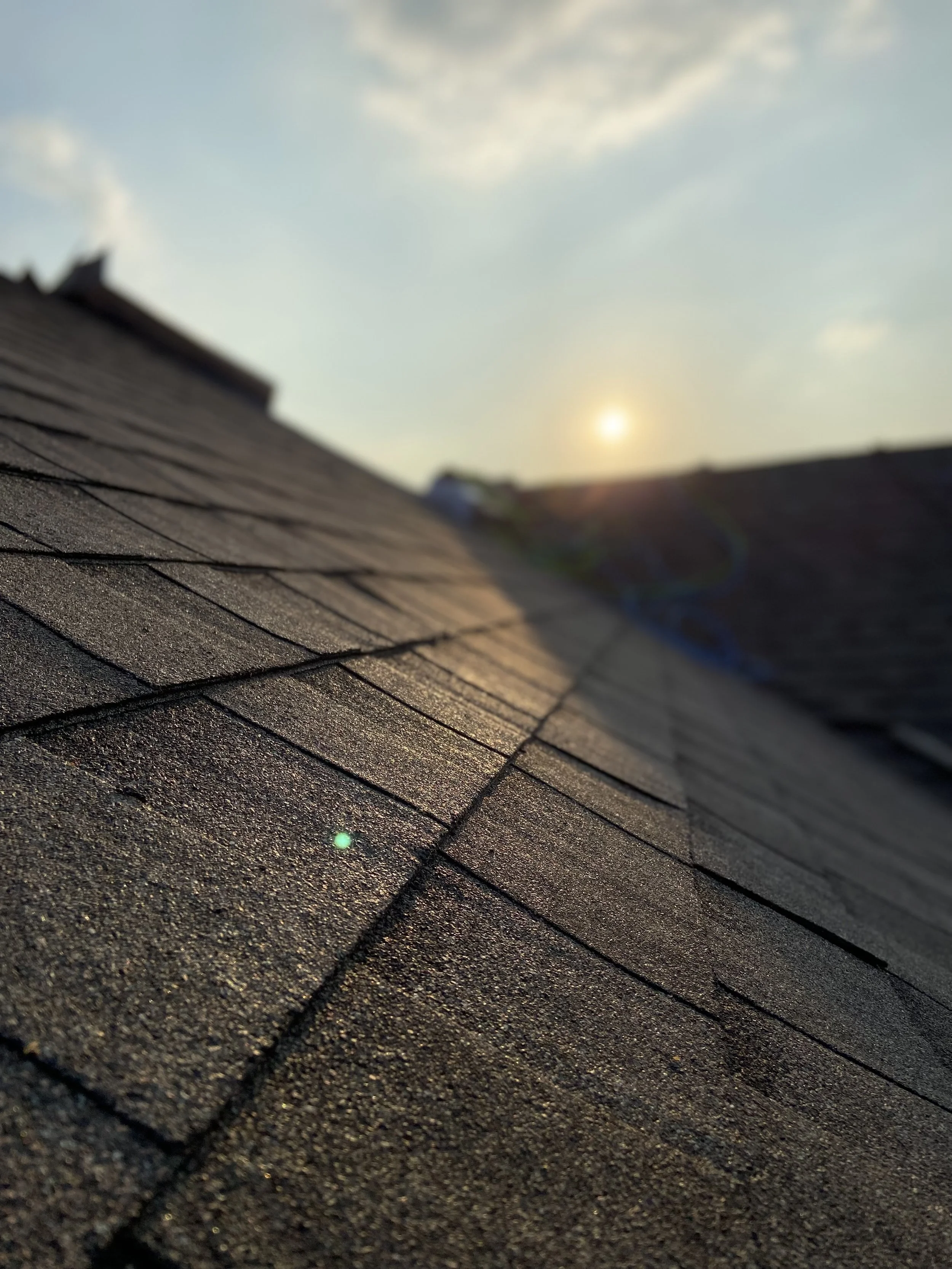 Close-up of a roof with asphalt shingles, with the sun setting in the background and a clear sky.