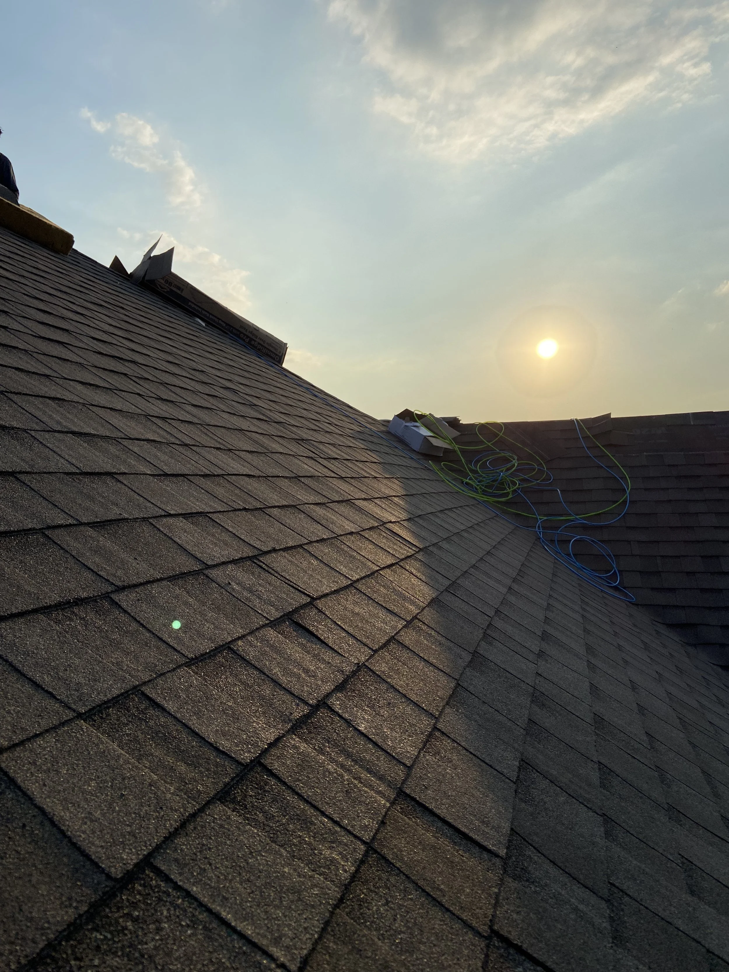 A roof under construction with shingles, electrical cables, and a vent, captured during sunset with a partly cloudy sky.