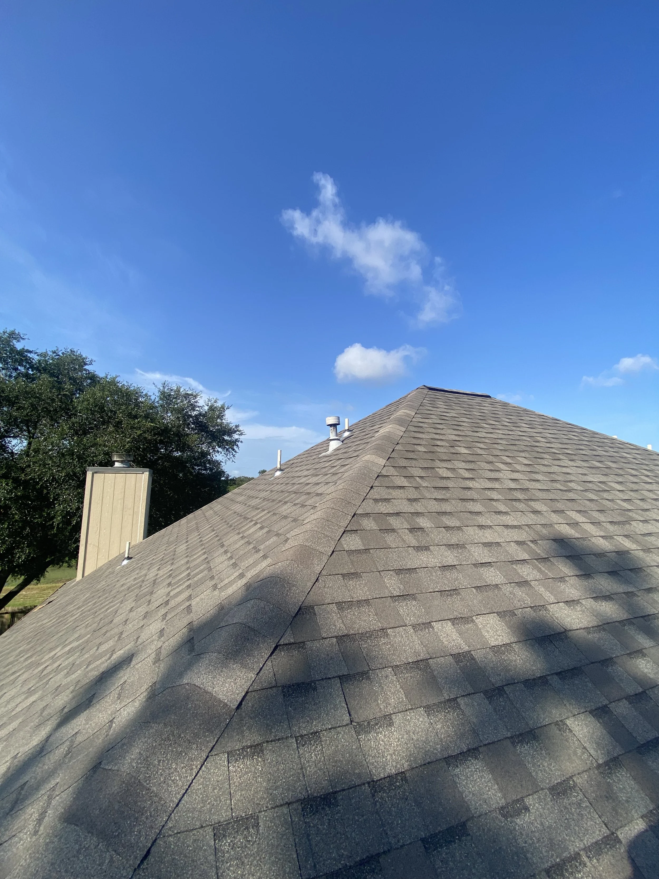 A rooftop with brown shingles under a blue sky with a few clouds, and trees in the background on a sunny day after a big storm.