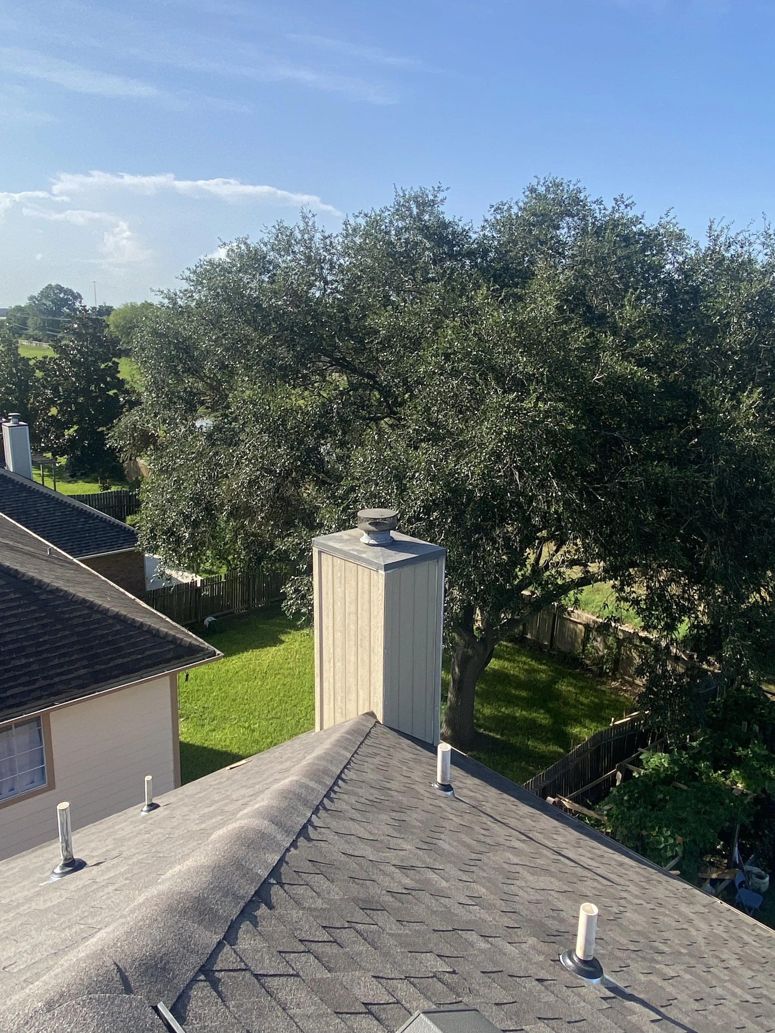 View of a residential backyard with trees and a rooftop with vents, under a blue sky with some clouds.