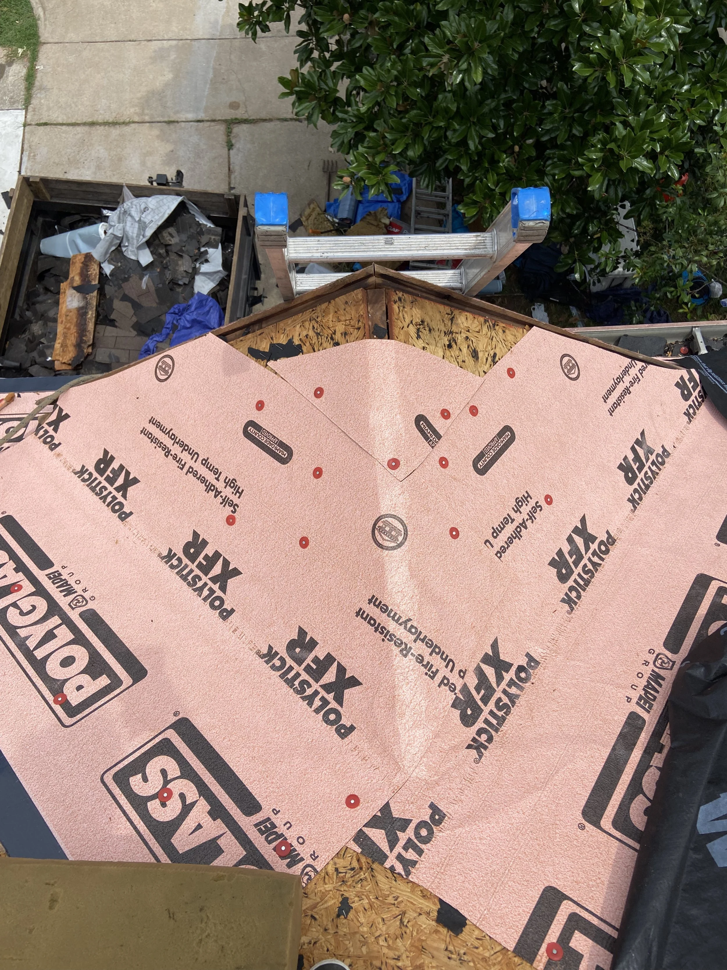 Roof under construction with pink high-temp roofing underlayment marked 'POLYGLASS XP3'. Ladder, construction tools, and debris visible on the roof, with a tree and some items on the ground below.