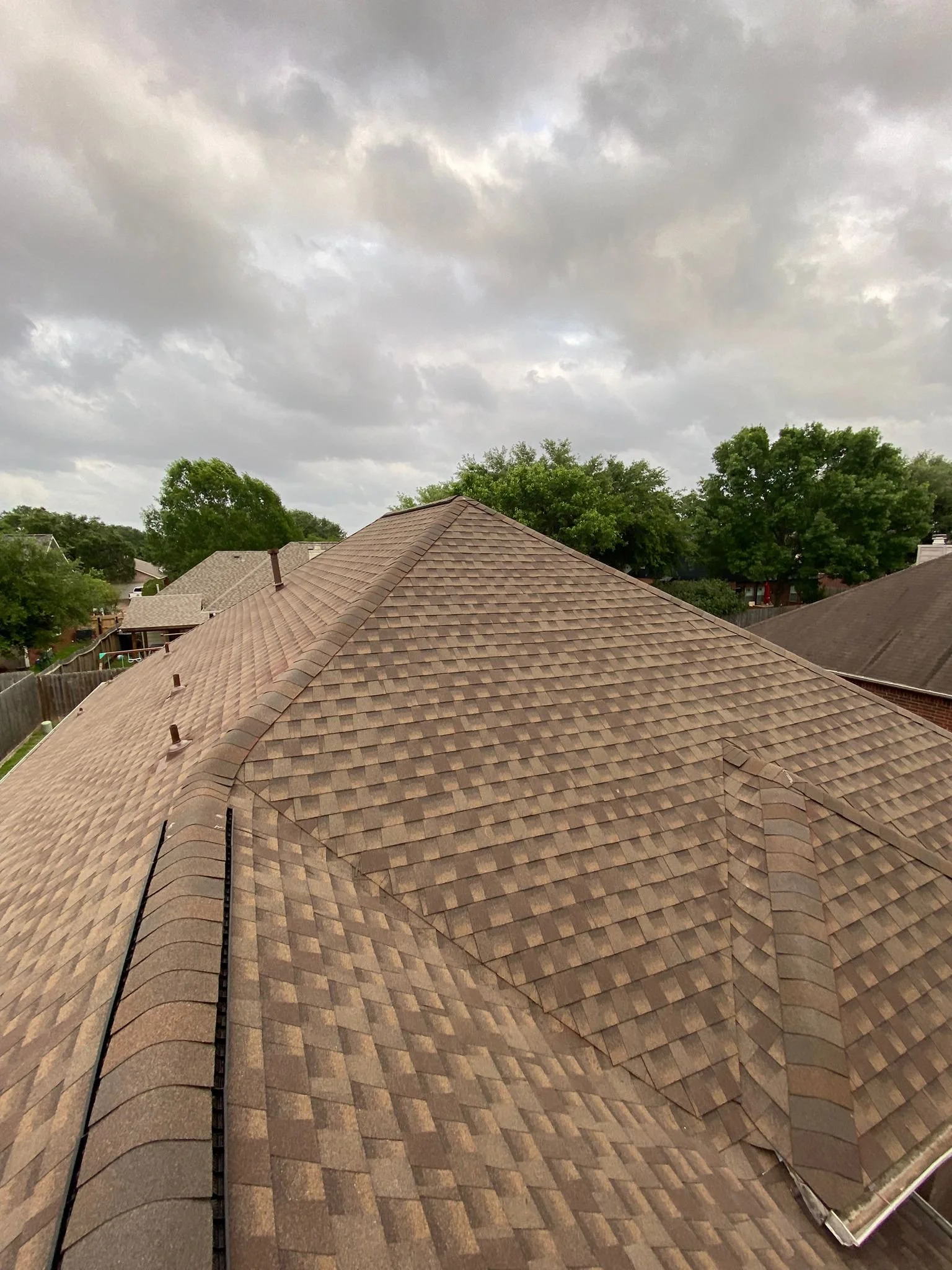 View of a house roof with brown shingles underneath a cloudy sky, surrounded by trees and neighboring houses.