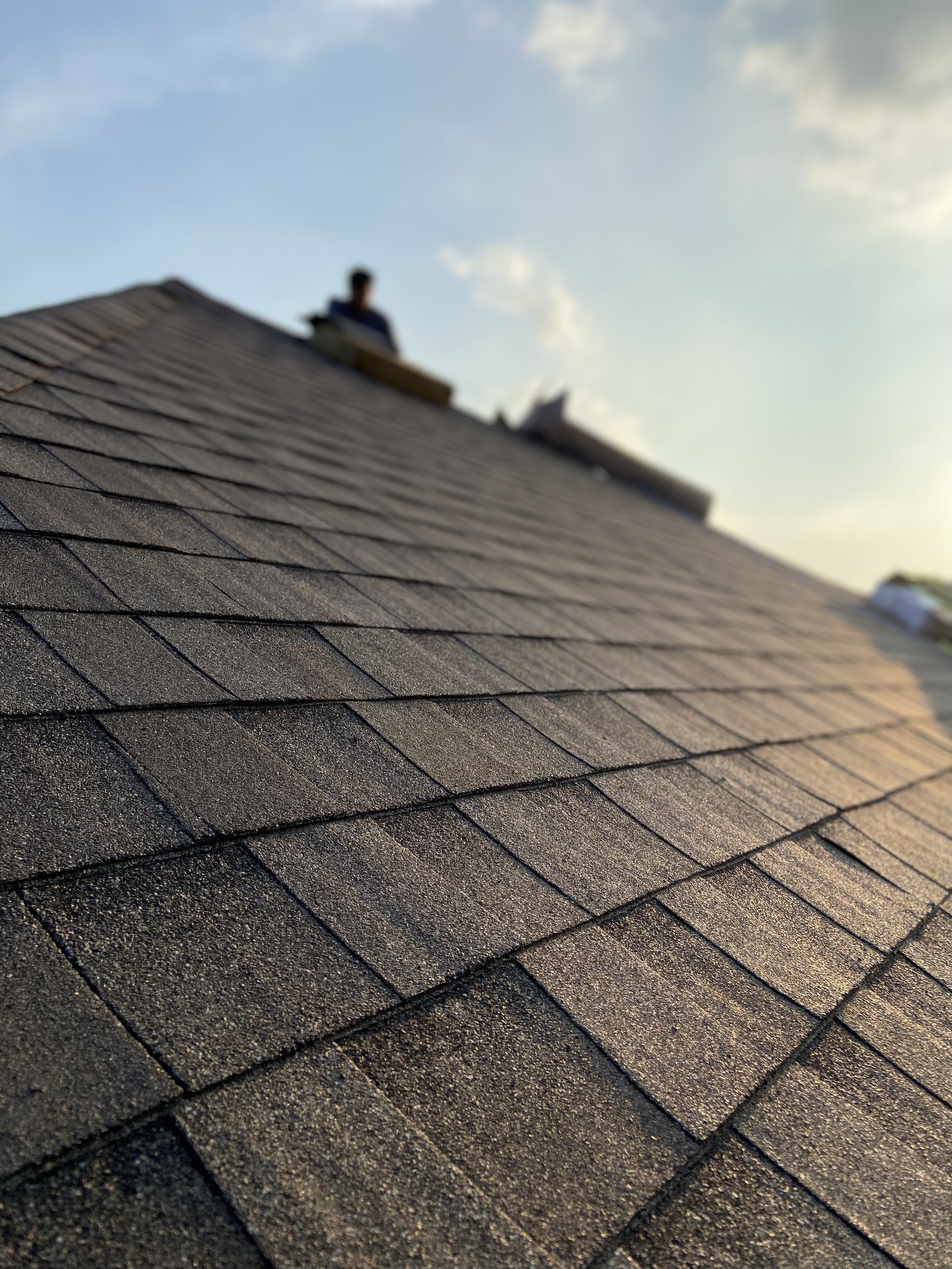 Close-up of a residential roof with brown asphalt shingles and a person sitting on top in the background under a partly cloudy sky.