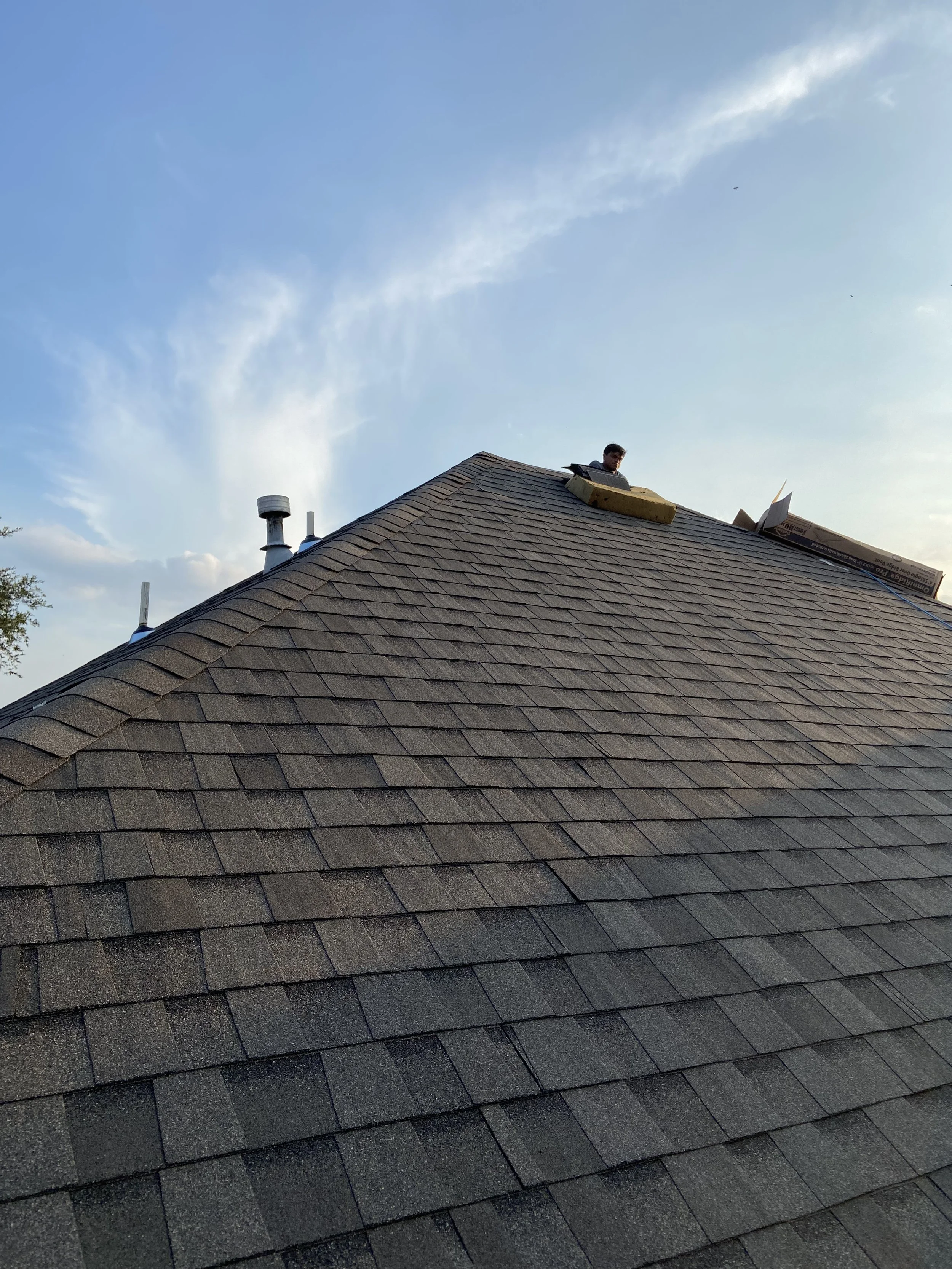 Person working on a sloped roof with shingles, looking down from above with a blue sky background.