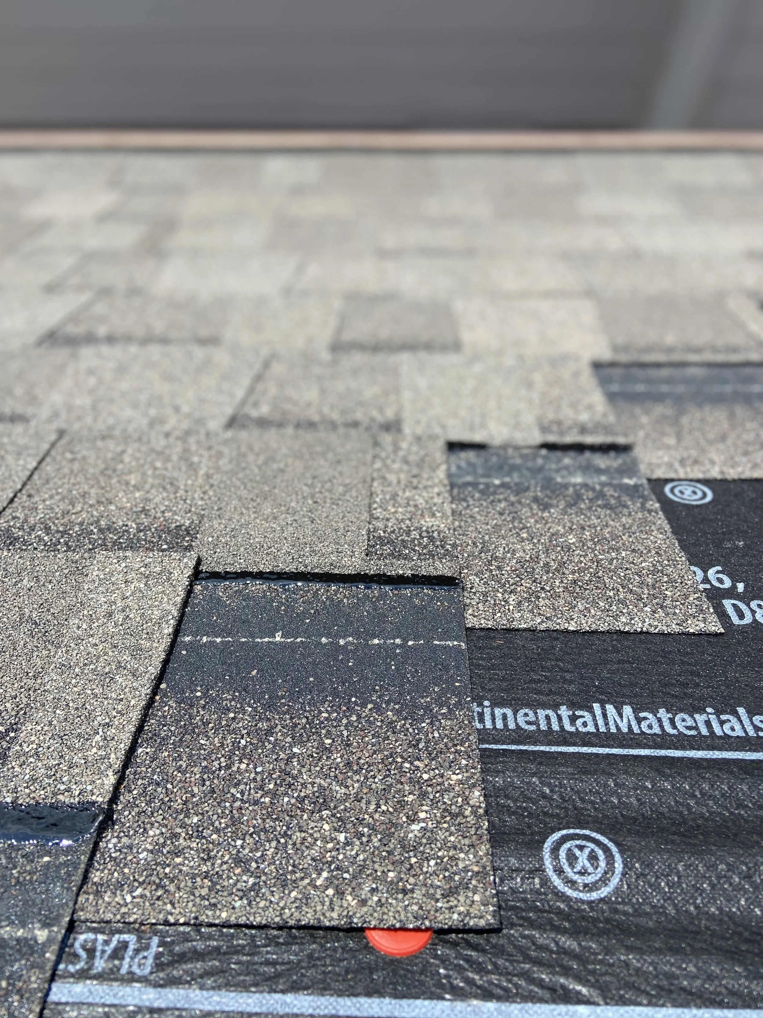 Close-up of asphalt roofing shingles in various shades of brown and black, with a black underlayment material showing partially at the bottom.