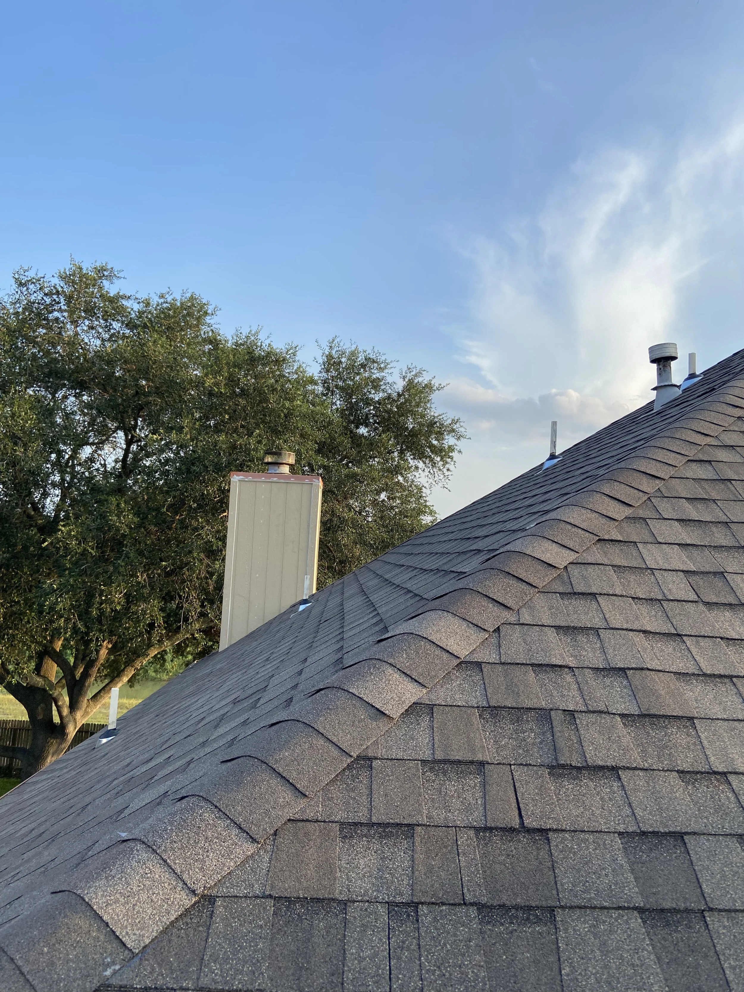 A rooftop with asphalt shingles, a large tree in the background, and a partly cloudy blue sky.