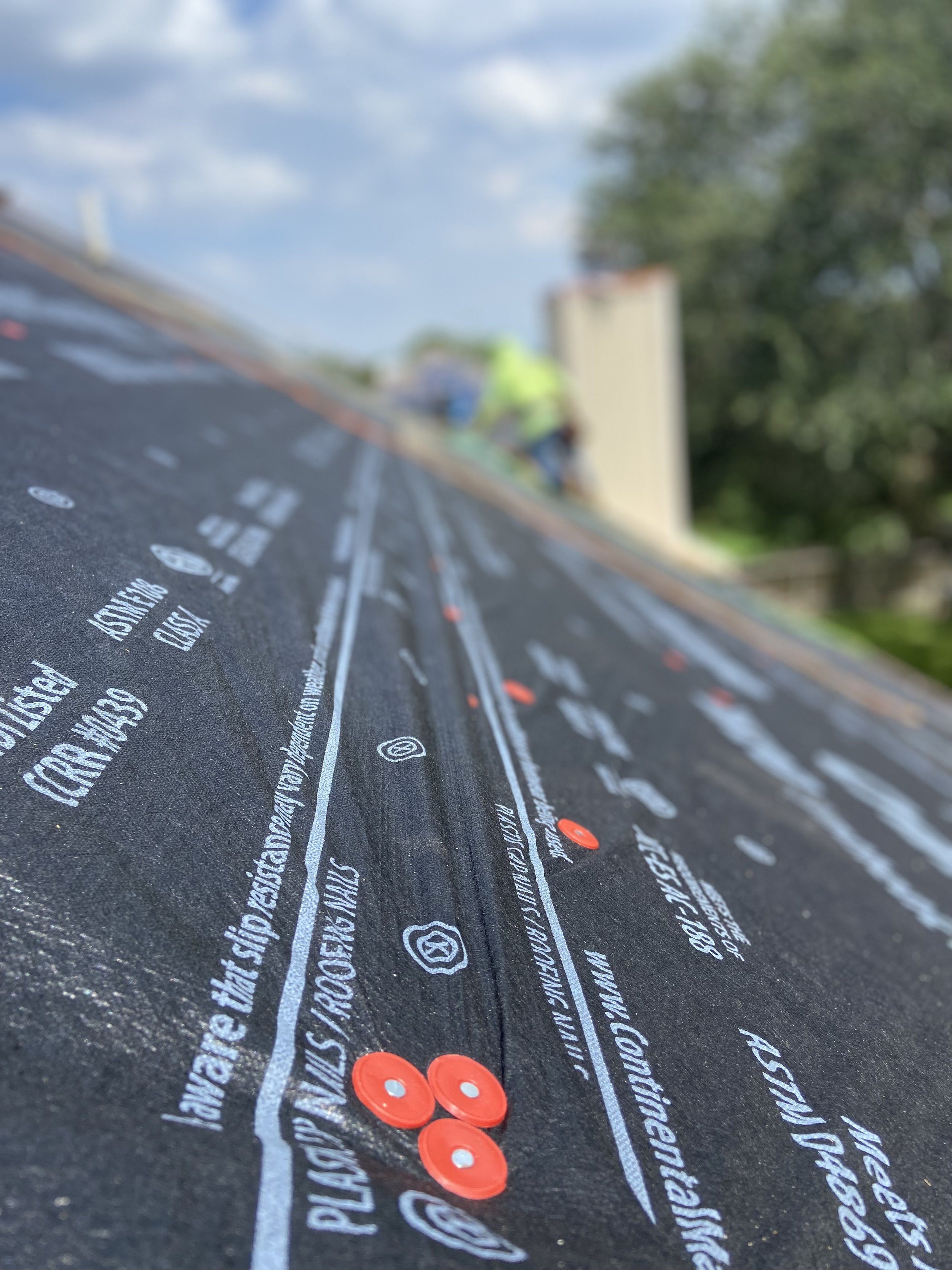 Close-up of black roofing underlayment with white printed text and red plastic caps, with a blurred background showing a person and house.