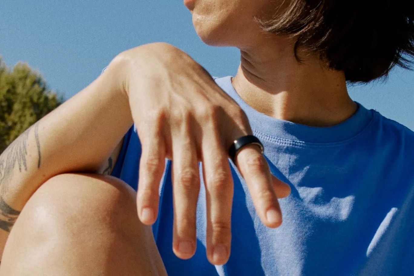 Close-up of a woman in a blue shirt showing her hand with a black ring, with trees and blue sky in the background.