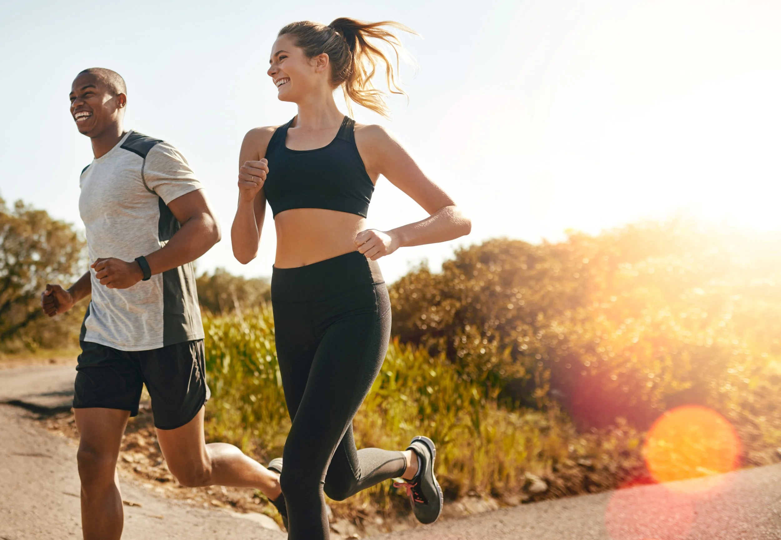 Two people running outdoors on a sunny day, a woman in a black sports bra and black leggings, a man in a light-colored t-shirt and black shorts, both smiling.