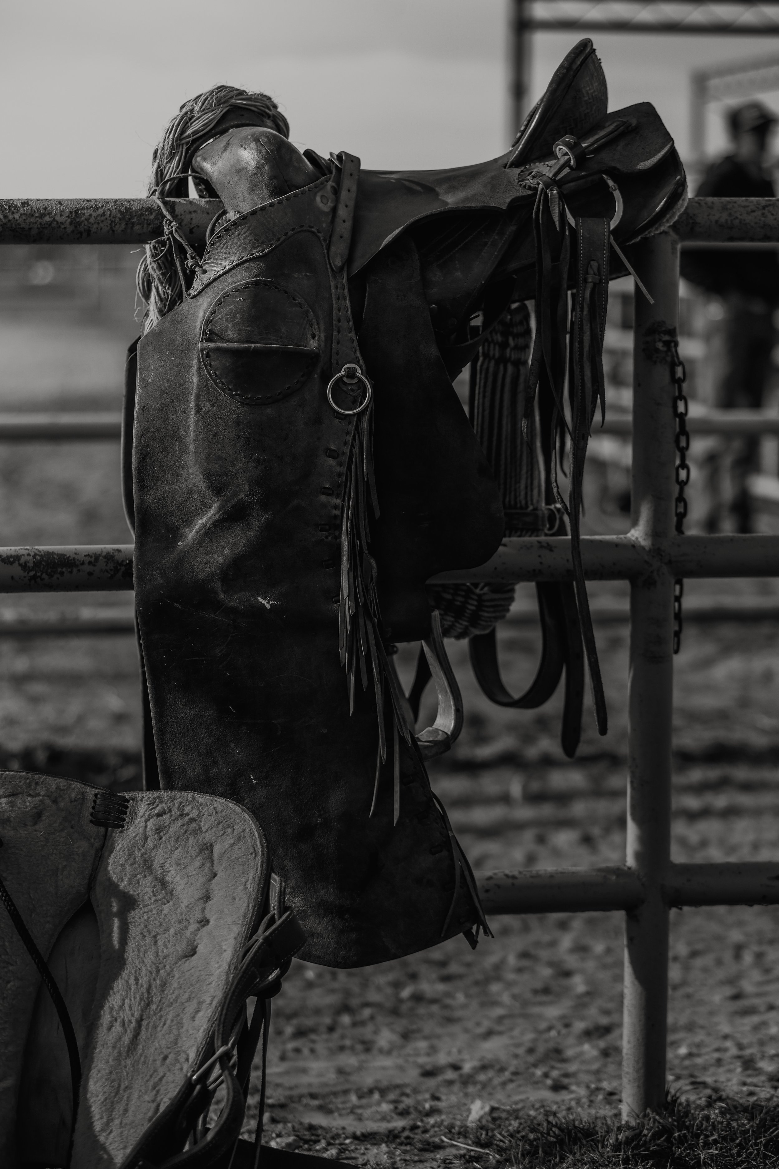A black and white image of a horse saddle hanging on a fence.