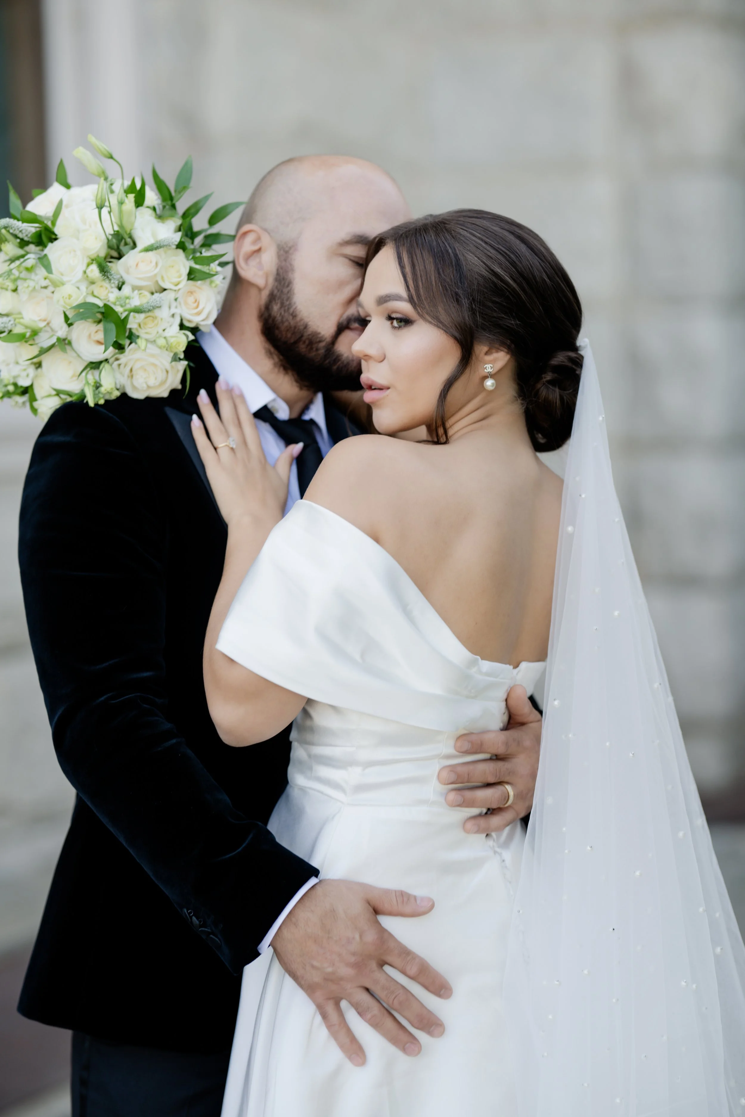 A bride and groom sharing an intimate moment, with the groom whispering in the bride's ear and holding her waist. The bride is wearing a white dress and pearl earrings, and the groom is dressed in a black suit, holding a large bouquet of white roses.