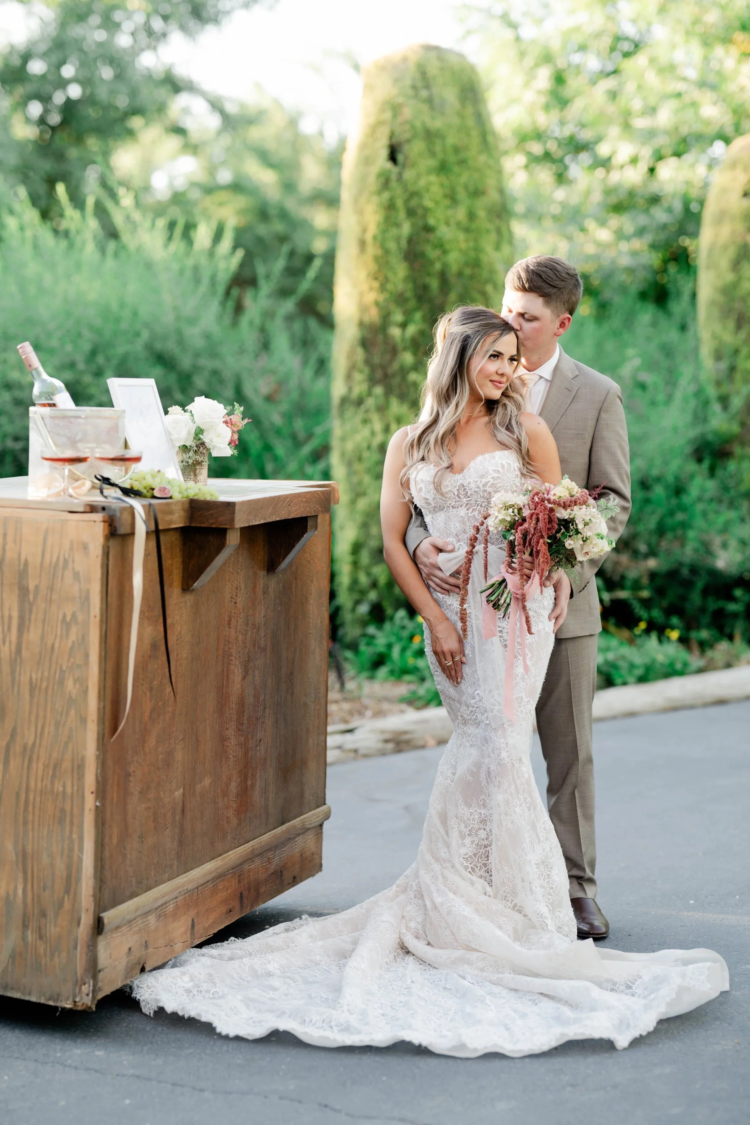 A newlywed couple standing outdoors near a wooden bar. The bride is holding a bouquet of flowers, and the groom is kissing her on the forehead. The setting is lush and green, suggesting a garden or park, with sunlight filtering through the trees.