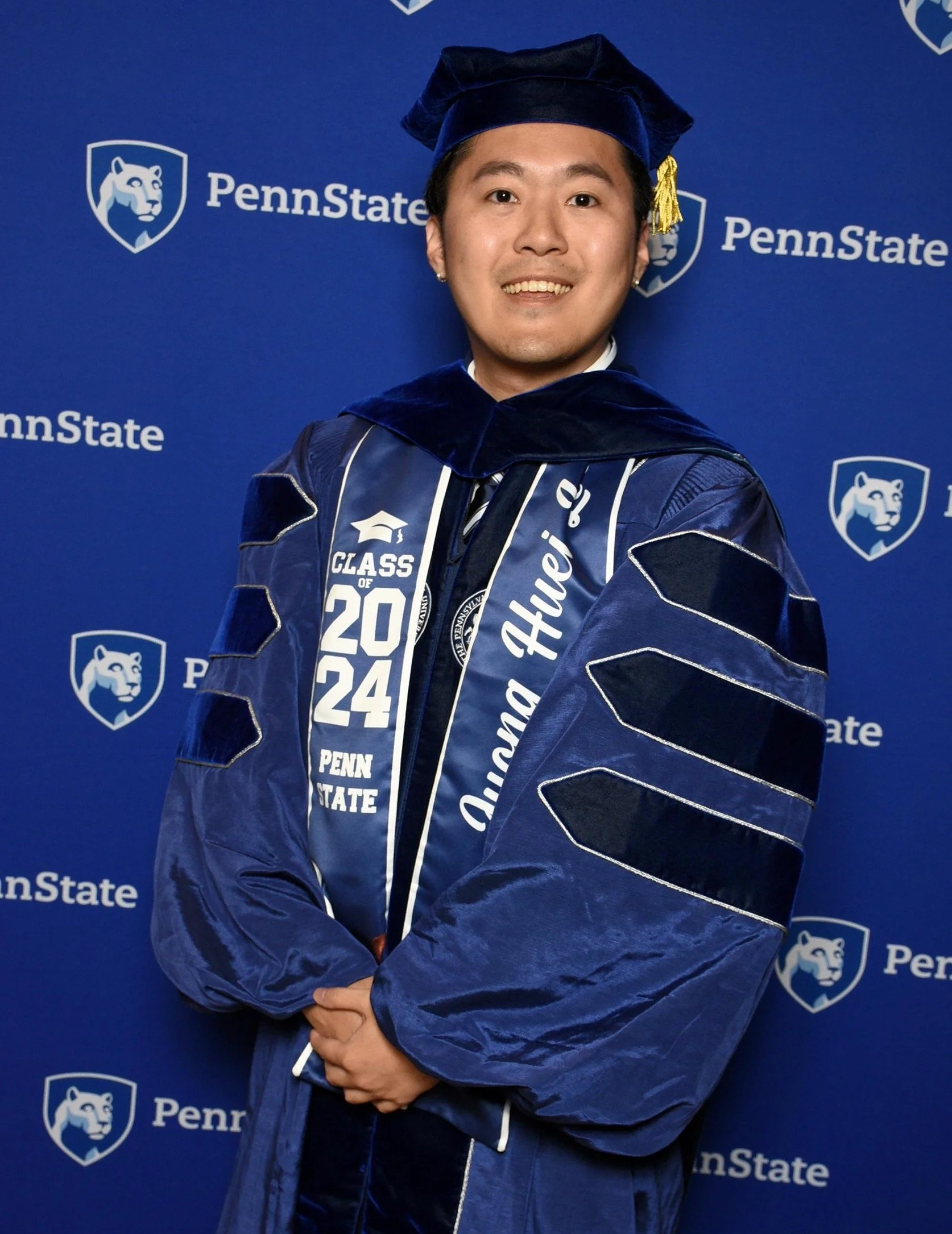 A young man in a cap and gown at a graduation ceremony, standing in front of a Penn State backdrop.
