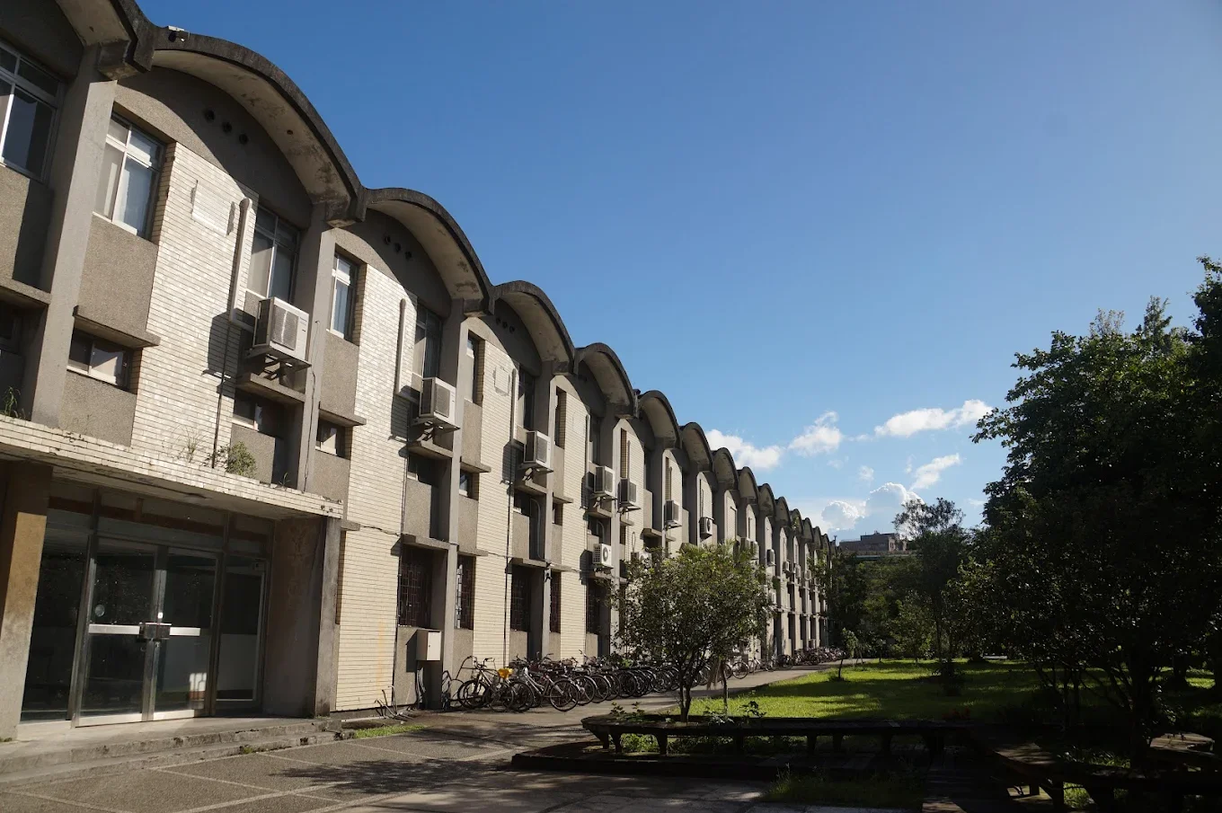 A multi-story apartment building with arched rooflines, air conditioning units, and bicycles parked outside on a sunny day with a clear blue sky and green trees.