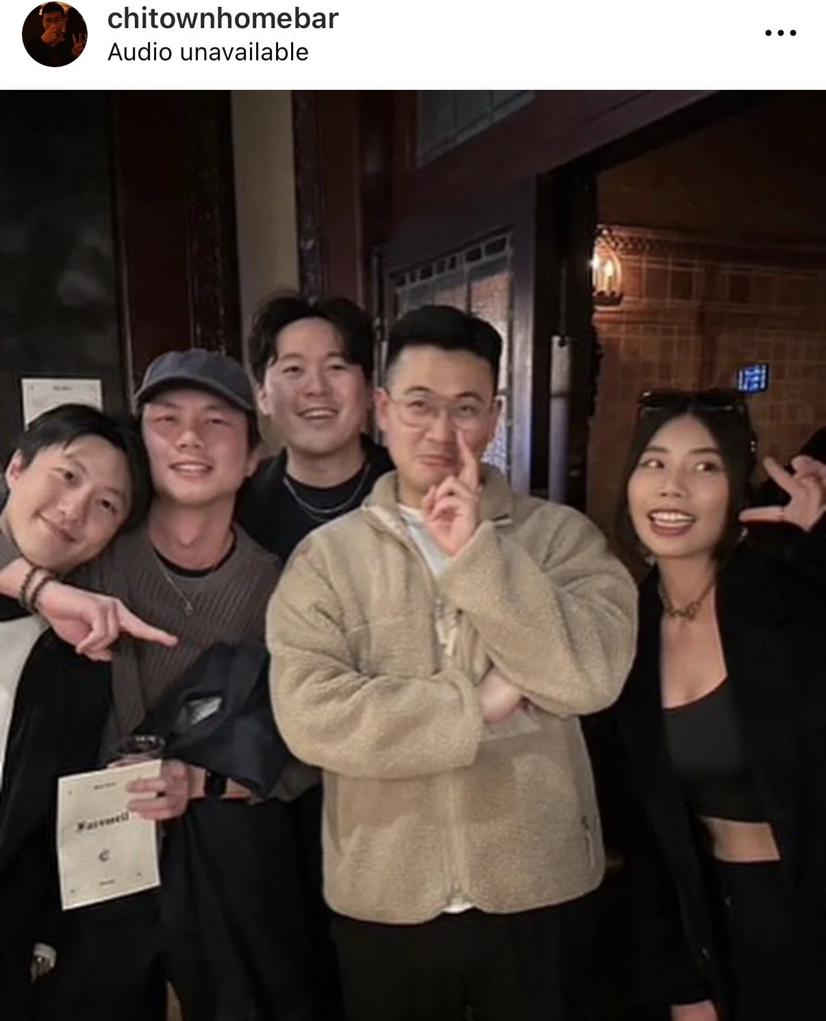 A group of six young adults posing together indoors, smiling and making playful gestures. The background has warm lighting and wooden decor.