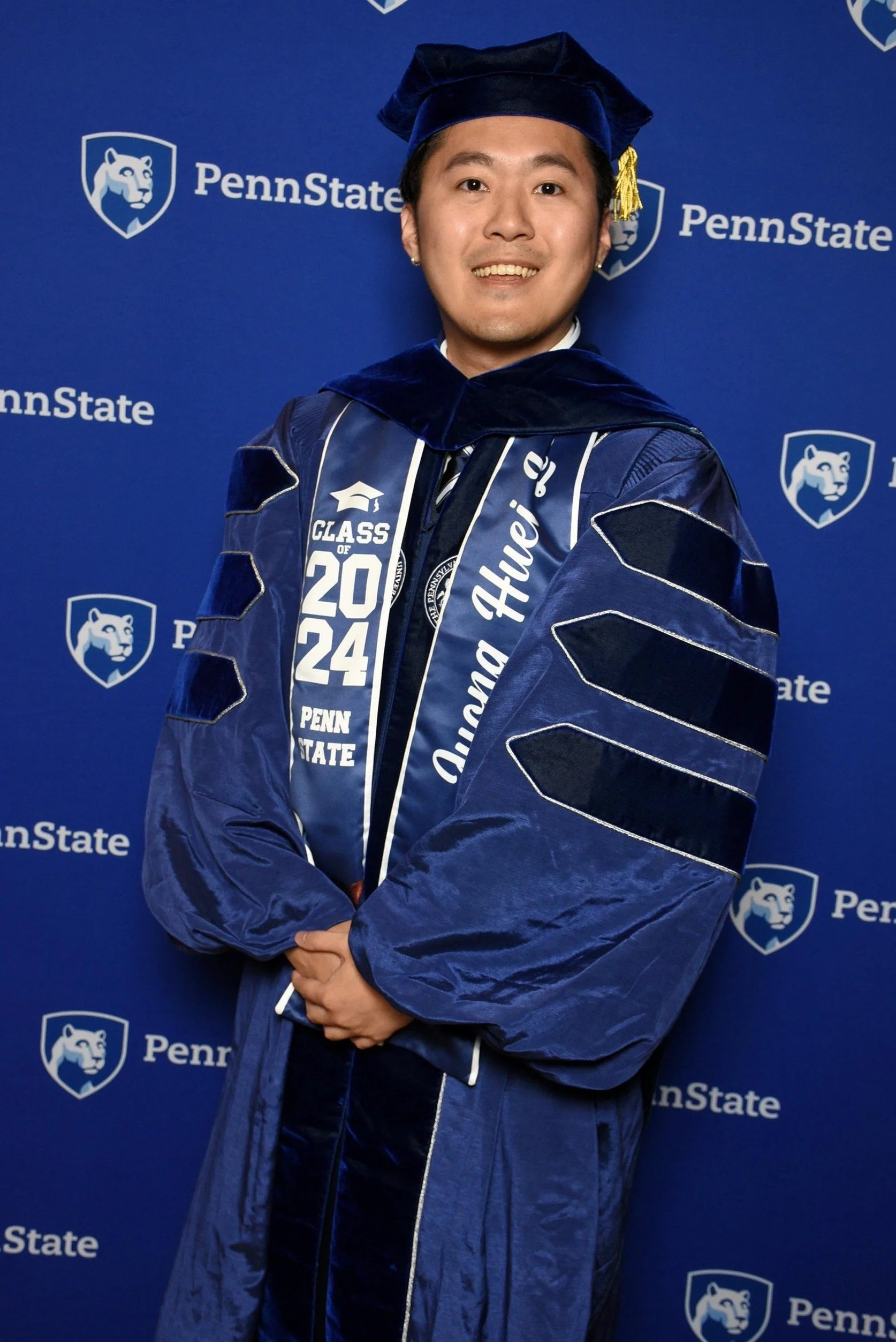 A young man in graduation robes posing in front of a blue Penn State backdrop, wearing a cap and gown with Penn State branding.