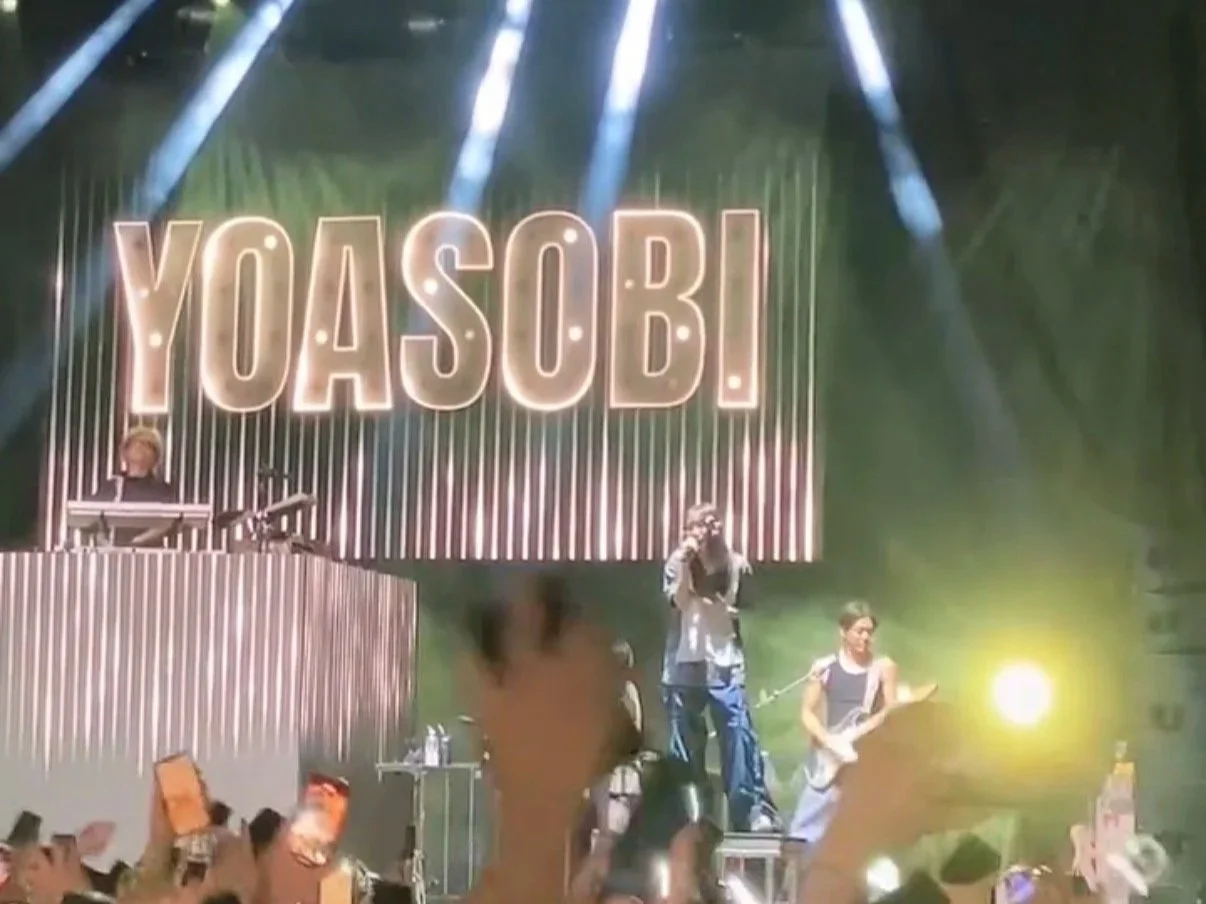 Musical performance on stage with large illuminated sign reading 'YOASOBI' in the background, band members playing instruments, bright stage lighting, audience with raised hands.