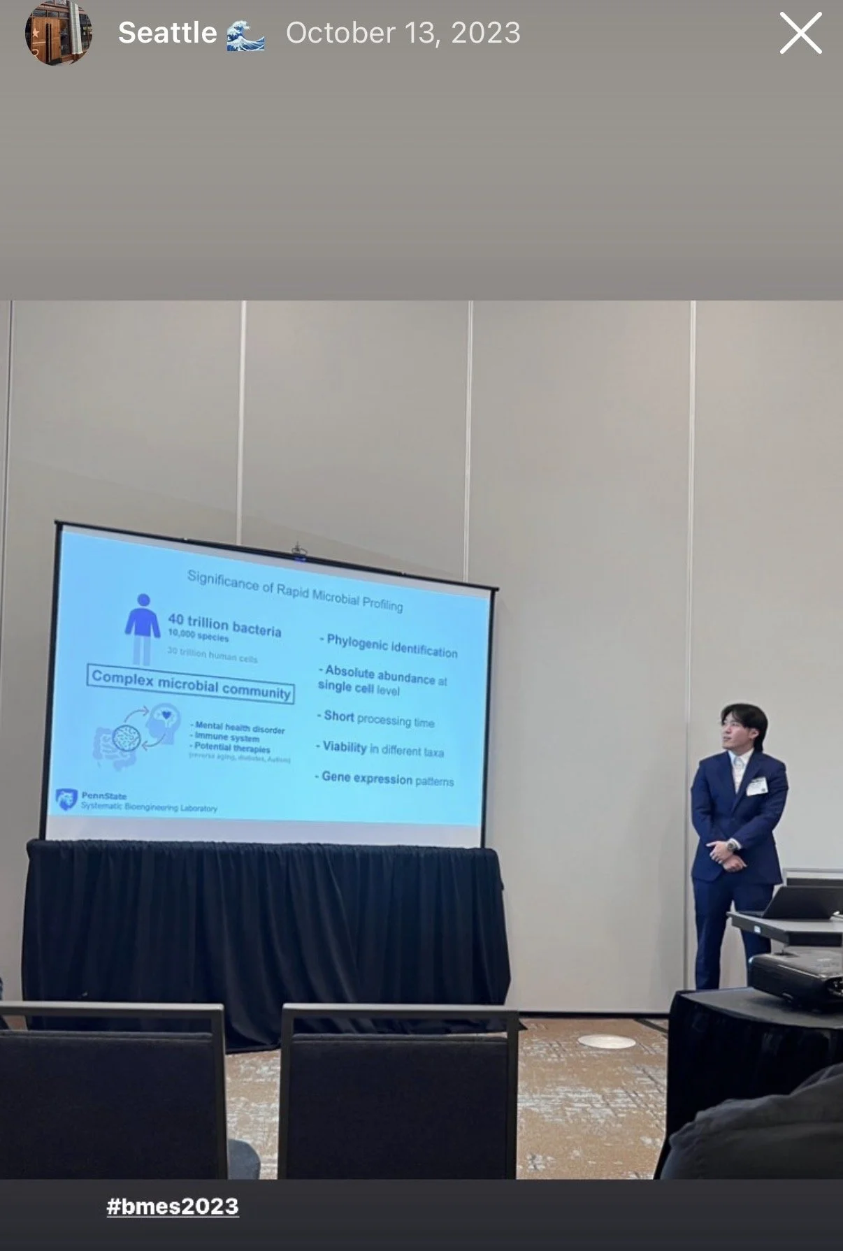 A man in a blue suit giving a presentation in a conference room. The presentation slide discusses the significance of rapid microbial profiling, mentioning statistics about bacteria and human cells, and topics like microbial community, phylogenetic identification, abundance, processing time, viability, and gene expression. The slide is from Penn State's Bioengineering Laboratory.