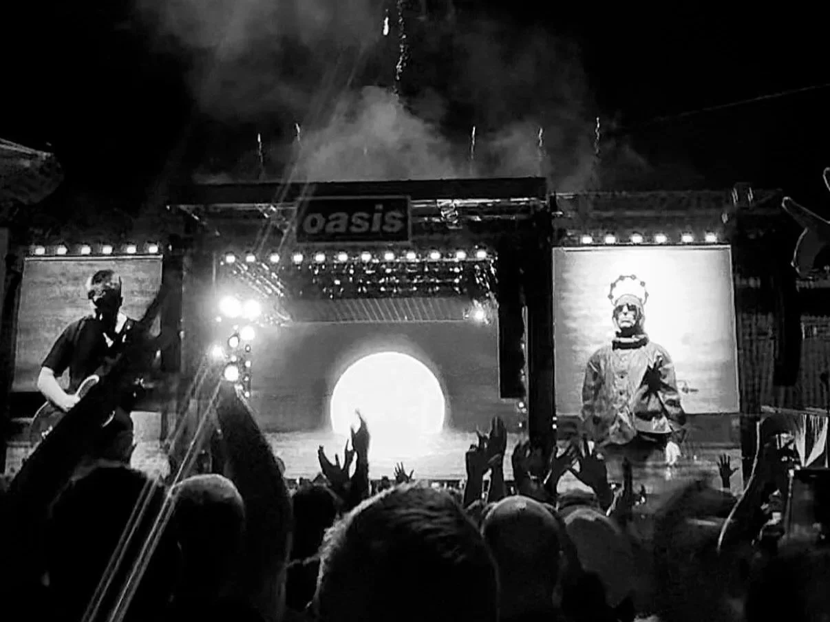 A black and white photograph of a concert with the stage lit up, showing a performer on the left with a guitar and a large screen on the right displaying an image of a man in traditional attire. The crowd in front has hands raised, and the background includes a sign with the word "Oasis."