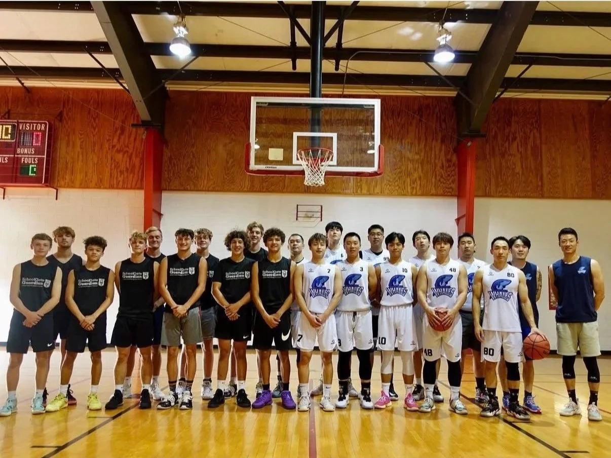 A group of male basketball players posing on a basketball court in front of a hoop, with some holding basketballs, in an indoor gymnasium.