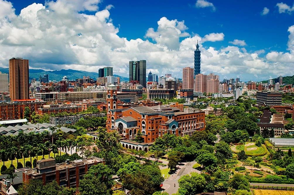 A city skyline featuring Taipei 101 tower, various high-rise buildings, and lush green parks under a bright blue sky with scattered white clouds.