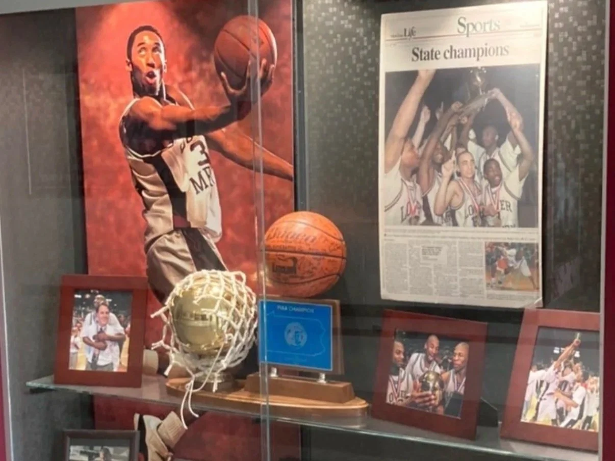 Display case with basketball memorabilia, including a framed photo of a basketball player, a small basketball hoop with a ball, a basketball, and photographs of basketball players celebrating a victory.