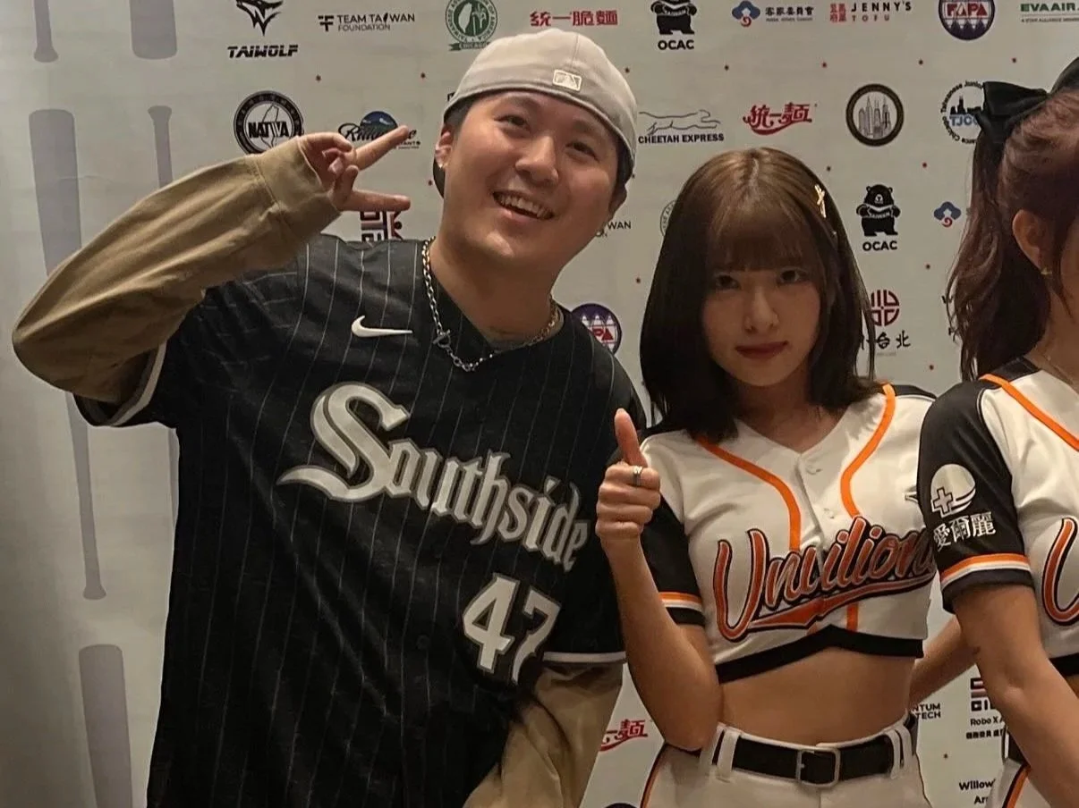 A young man and a woman in a sports jersey posing together at an event, smiling and making peace and thumbs-up gestures.