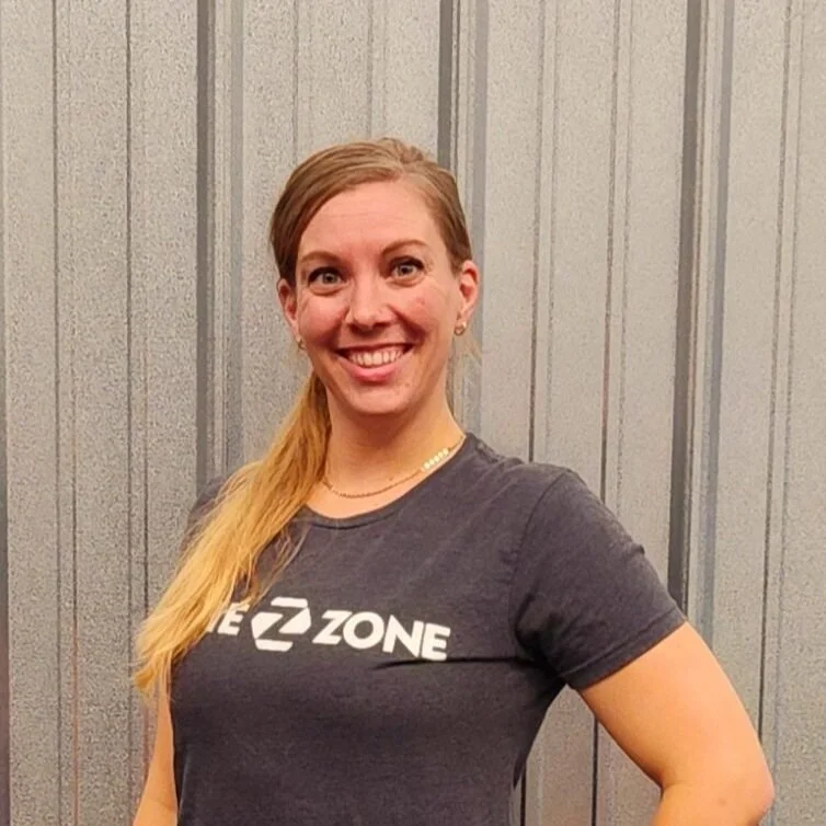 Smiling woman with long blonde hair, wearing a dark T-shirt with a logo, standing against a gray, striped wall.