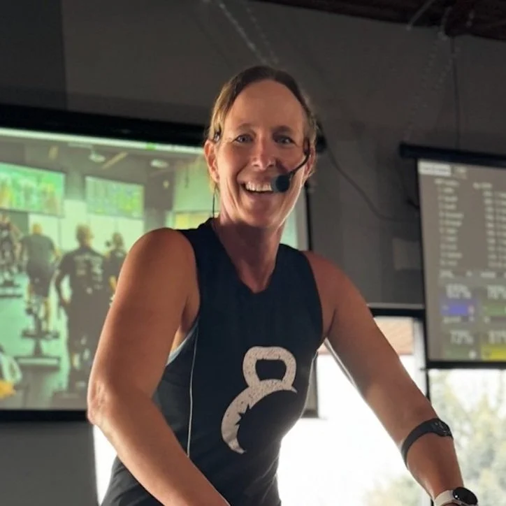 A woman wearing a sleeveless athletic top with a kettlebell logo, smiling and using a headset microphone during a fitness class at a gym, with fitness screens in the background.