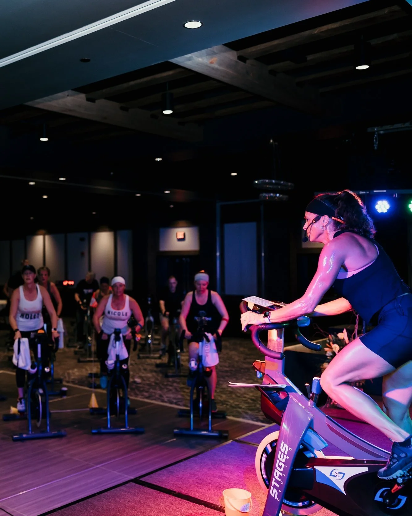 Indoor cycling class with a female instructor leading a workout, participants wearing workout clothes and headbands, dim lighting, colorful stage lights.