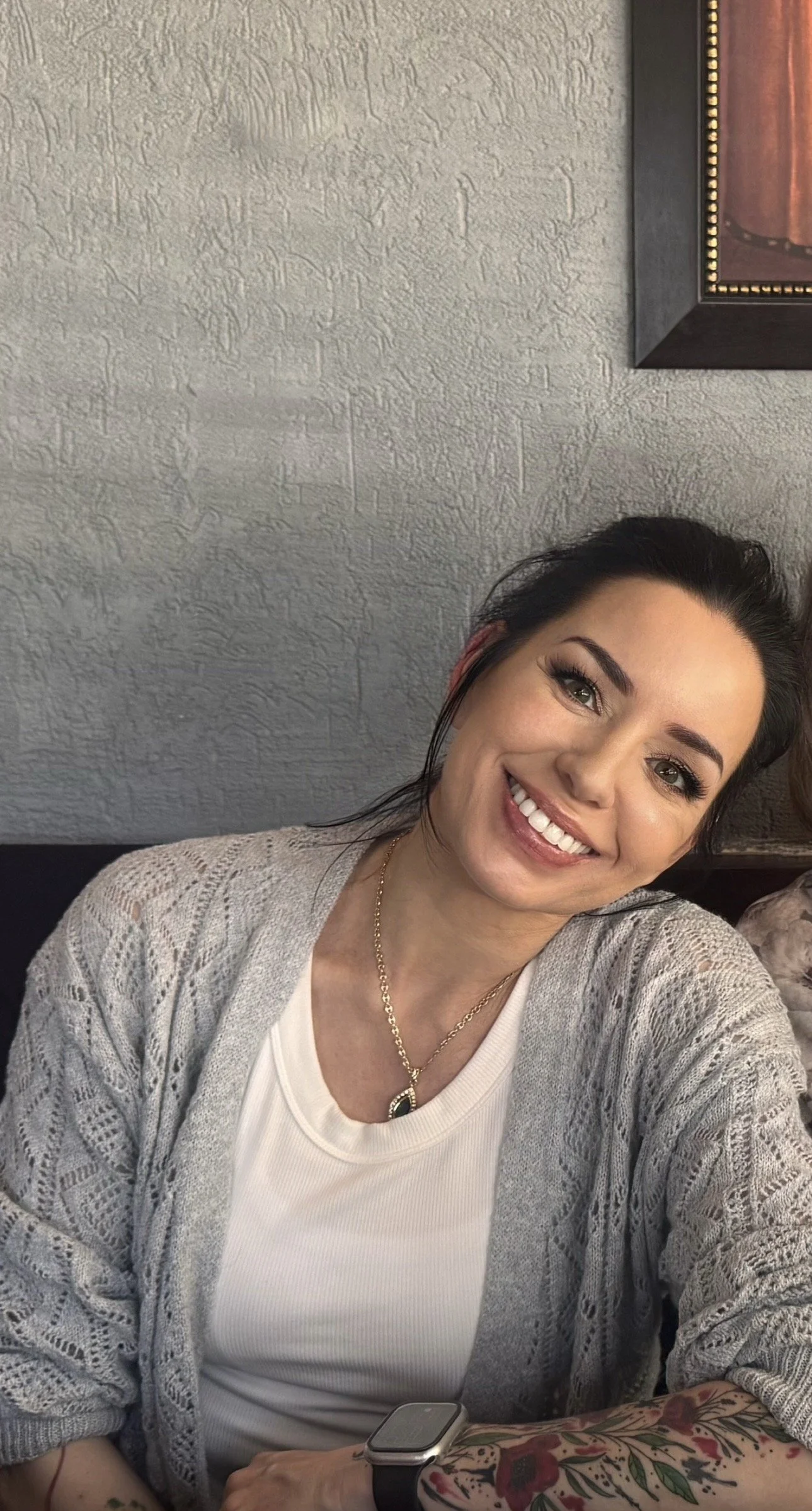 A smiling woman with dark hair, wearing a white top, gray knitted cardigan, and a gold necklace, taking a selfie indoors, with a textured gray wall and a framed picture in the background.