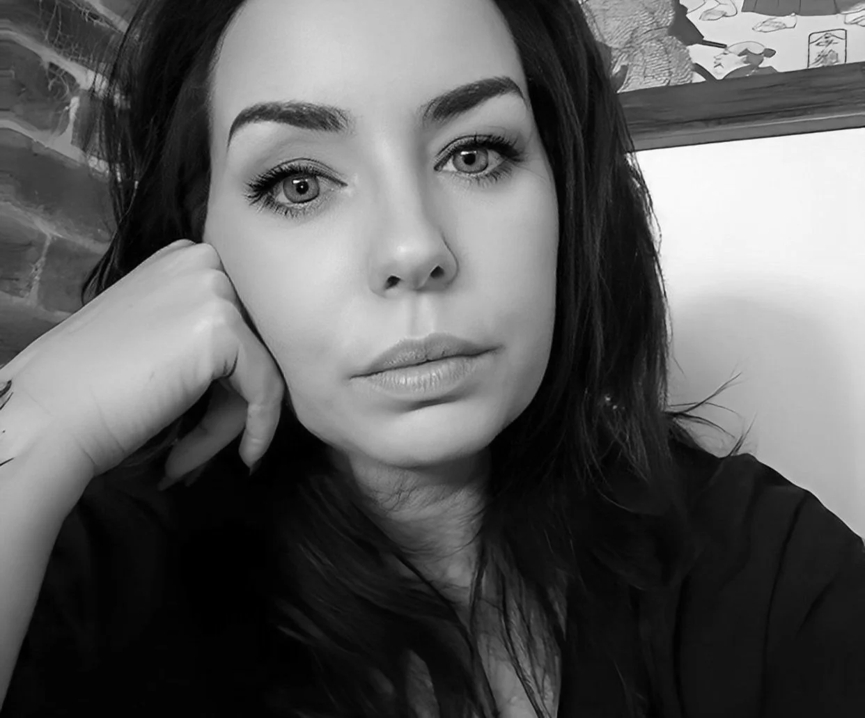Black and white portrait of a woman with dark hair, resting her face on her hand, looking directly at the camera with a neutral expression. Background includes brick wall and artwork.
