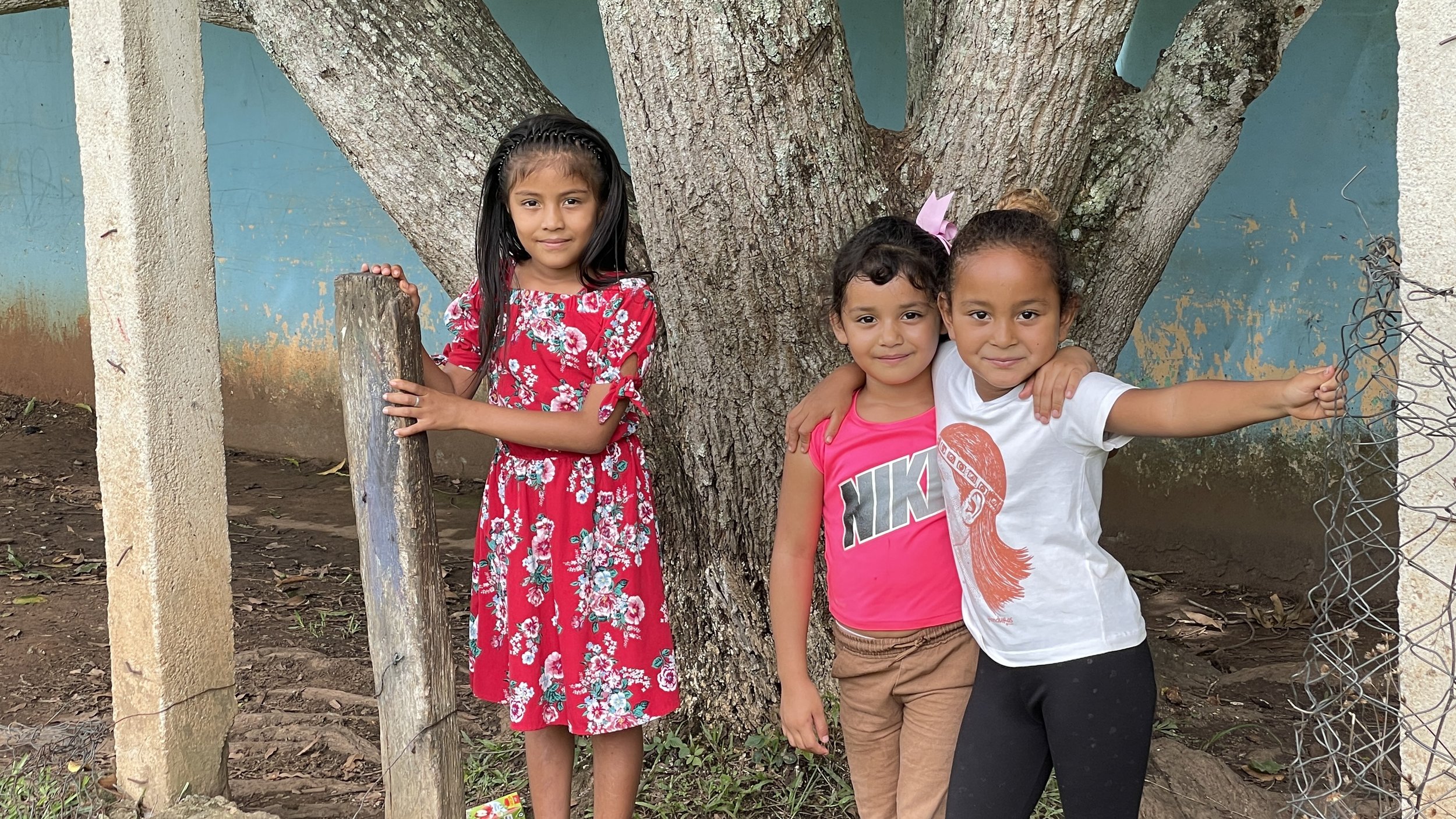 Three young girls standing outdoors in front of a large tree, with a blue and beige wall in the background. One girl wears a red floral dress, another a pink Nike shirt, and the third a white shirt with a graphic. One girl has her arm around another, smiling at the camera.