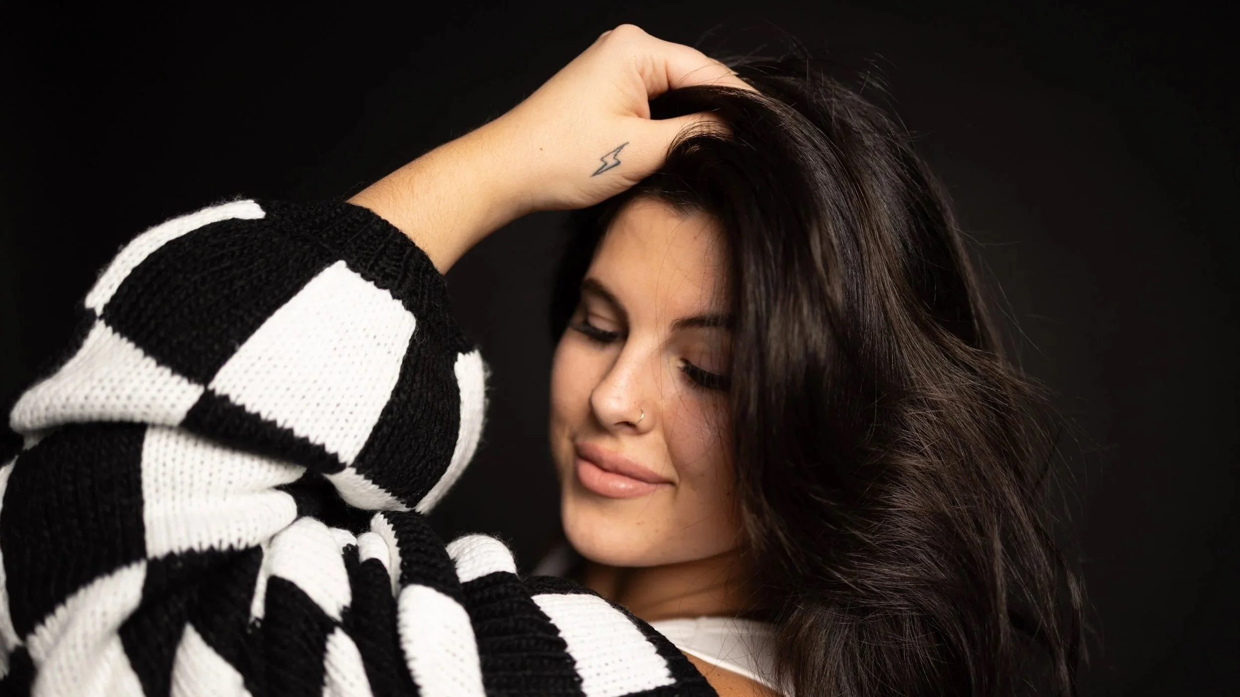 A woman with dark, wavy hair and a nose piercing, smiling subtly with eyes closed, holding her hair back with her hand, which has a small lightning tattoo, against a black background. She is wearing a black and white striped sweater.