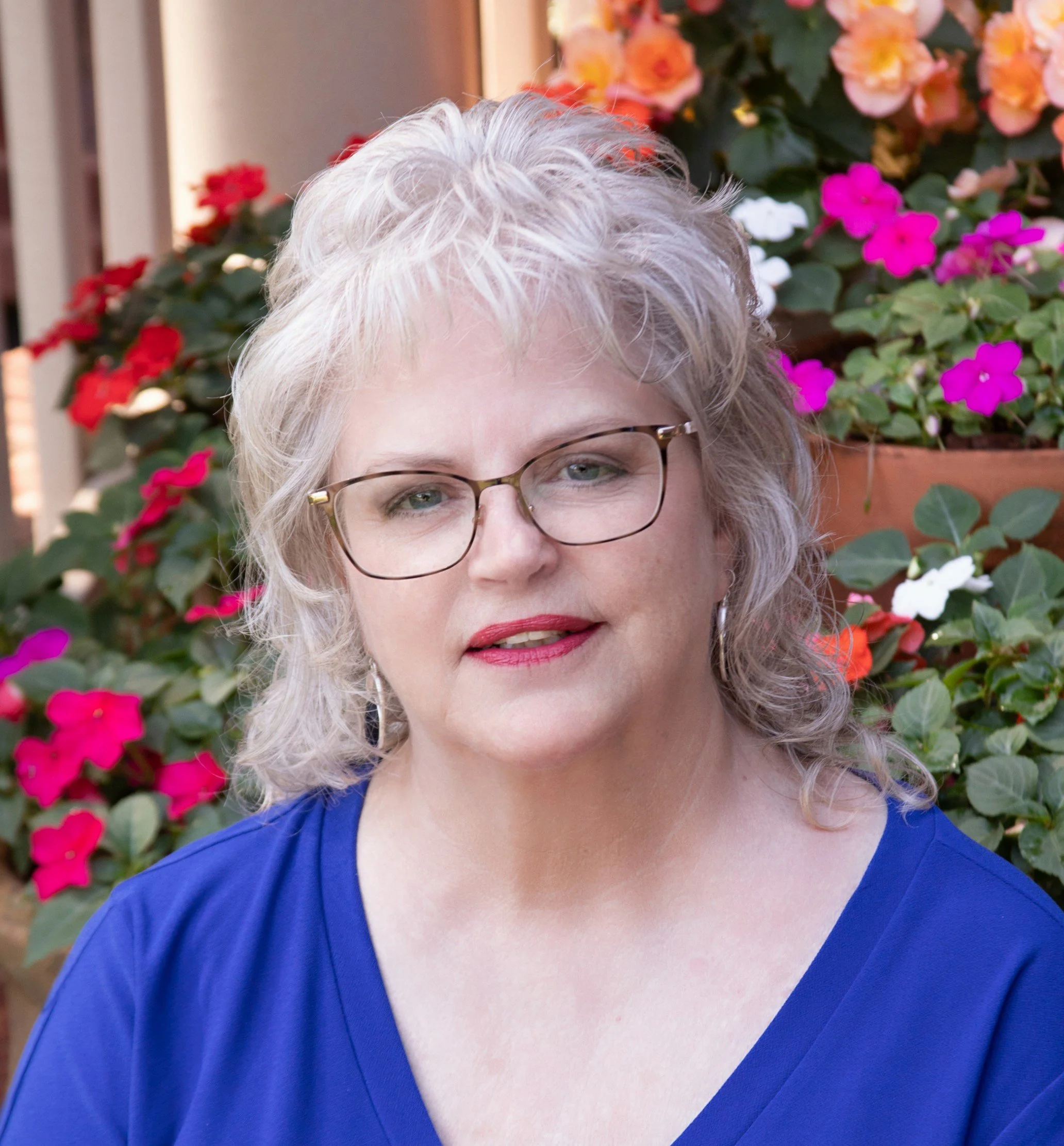 Image of Nancy Ruffner, upper torso and headshot. Smiling. Wearing glasses. Graying hair. Sitting on porch steps with colorful flowers in backround.