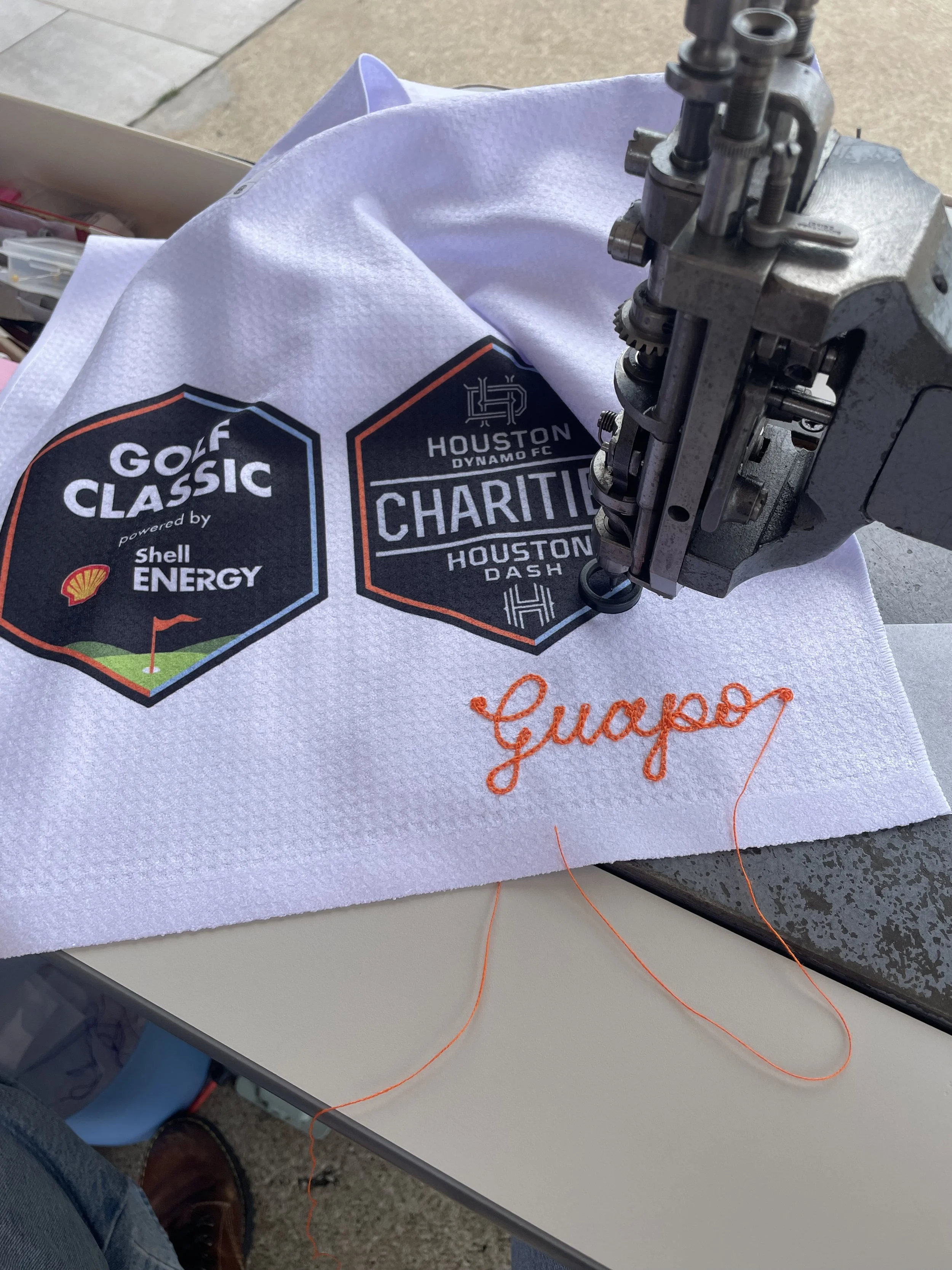 A machine chainstitch embroidery process creating a name with the text 'Guapo' on a white golf towel. The fabric features logos for Houston Dynamo FC, Golf Classic powered by Shell Energy, and Houston Dash. Double Vision was on-site to provide live e