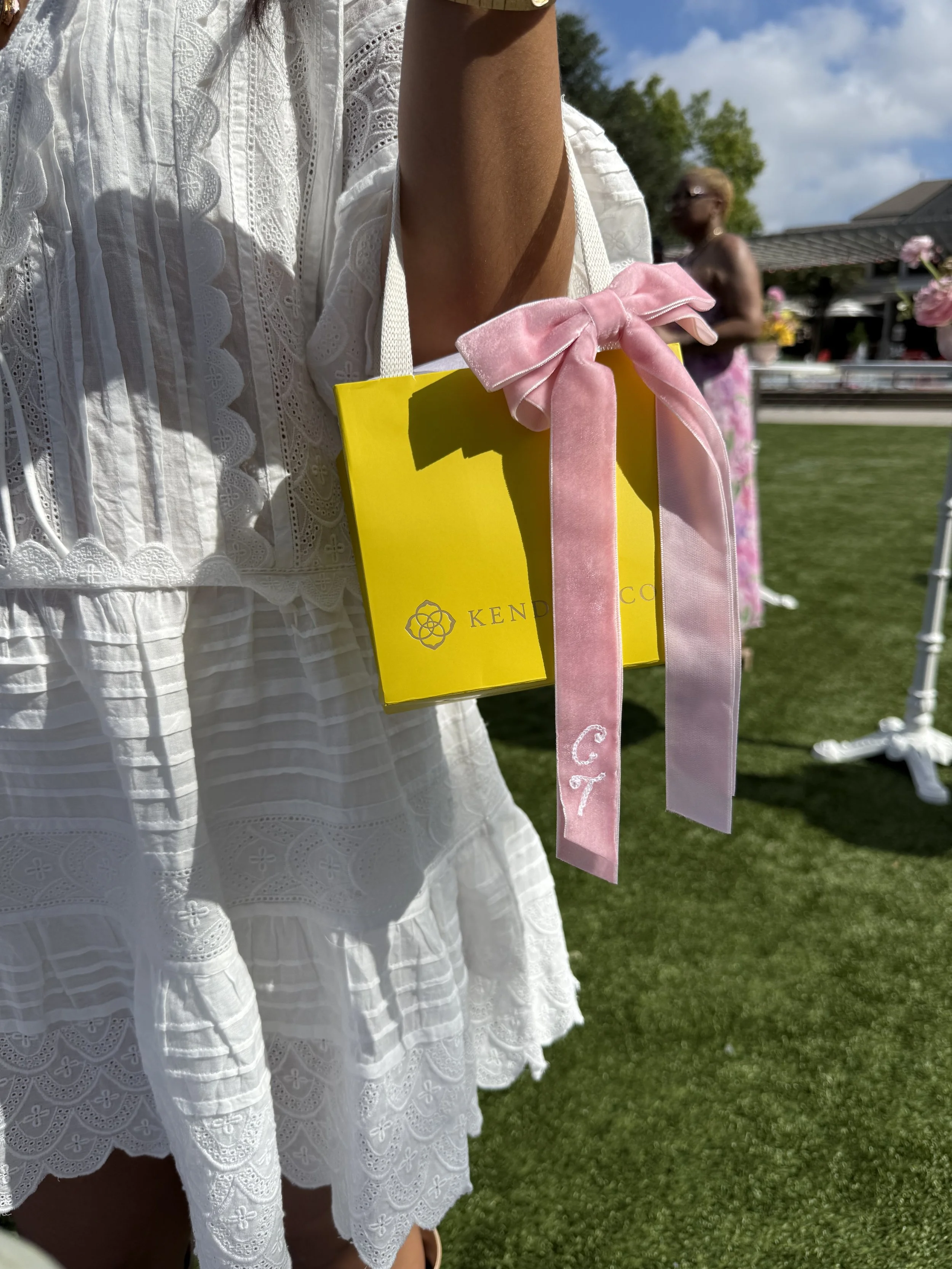 A woman in a white lace dress holding a yellow box with a pink ribbon with chainstitch embroidered initials, with another woman in a floral dress in the background on a grassy outdoor setting. Double Vision was in The Woodlands with Kendra Scott to p
